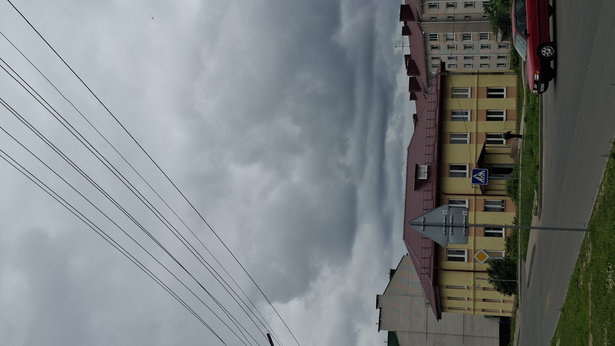 The image depicts a street scene in Smalyavichy, Belarus, under a heavy, overcast sky. In the foreground, a two-lane asphalt road curves to the left, with a grassy median and sidewalk. A red sedan is driving on the road, heading towards the right of the frame. To the left of the road is a yellow building with a burgundy roof, featuring multiple windows and an entrance. In front of this building, a pedestrian crossing sign and a yield sign are visible on a metal pole. A person in dark clothing is walking on the sidewalk in front of the yellow building. Further in the background, to the right of the yellow building, a multi-story residential building with a burgundy roof and numerous windows is situated. The sky is filled with dark, dense clouds, suggesting impending rain. Overhead, several power lines are strung across the frame, contributing to the urban atmosphere. The overall environment is that of a residential street in a town or city.