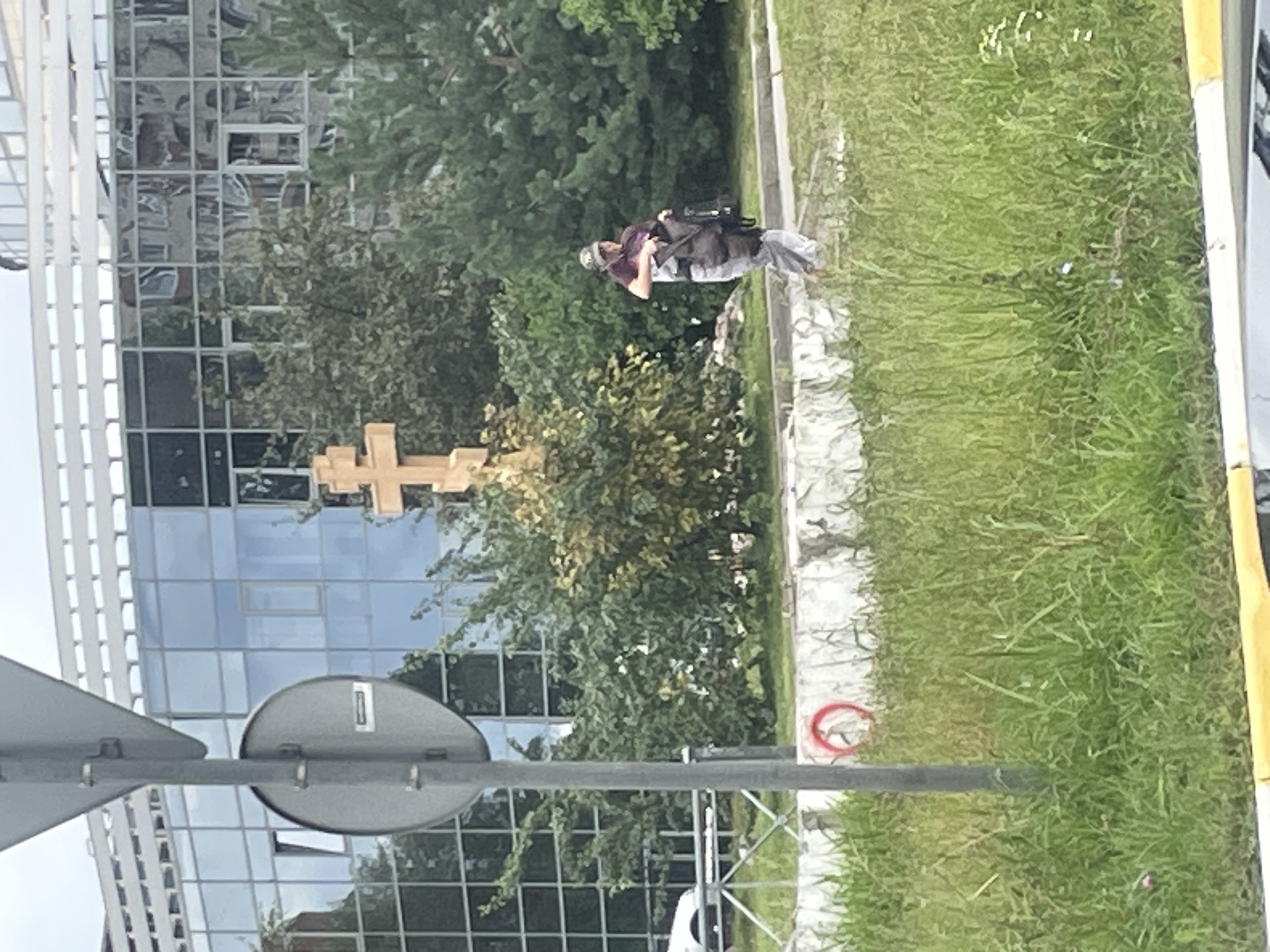 The image depicts an outdoor scene in Saint Petersburg, Russia. In the foreground, a tall, gray metal pole with a triangular sign and a circular sign attached is positioned on the left side of the frame. The circular sign appears to have some text or markings on it. Behind the pole, there is a patch of green grass with some weeds and a small pink flower visible. Further back, a white concrete barrier runs horizontally across the middle ground. A red circular object with a white symbol inside is affixed to this barrier.
In the background, a person wearing a patterned cap, a maroon t-shirt, and light-colored pants is seen walking. They are carrying a dark bag or backpack over their shoulder. To the left of the person, a large, wooden Orthodox Christian cross stands prominently. Behind the cross and the person, there is lush greenery, including trees and bushes. The very back of the image features a modern building with a glass facade. Reflections of buildings and the sky are visible in the glass. The overall environment suggests an urban setting with a mix of natural elements and architecture.