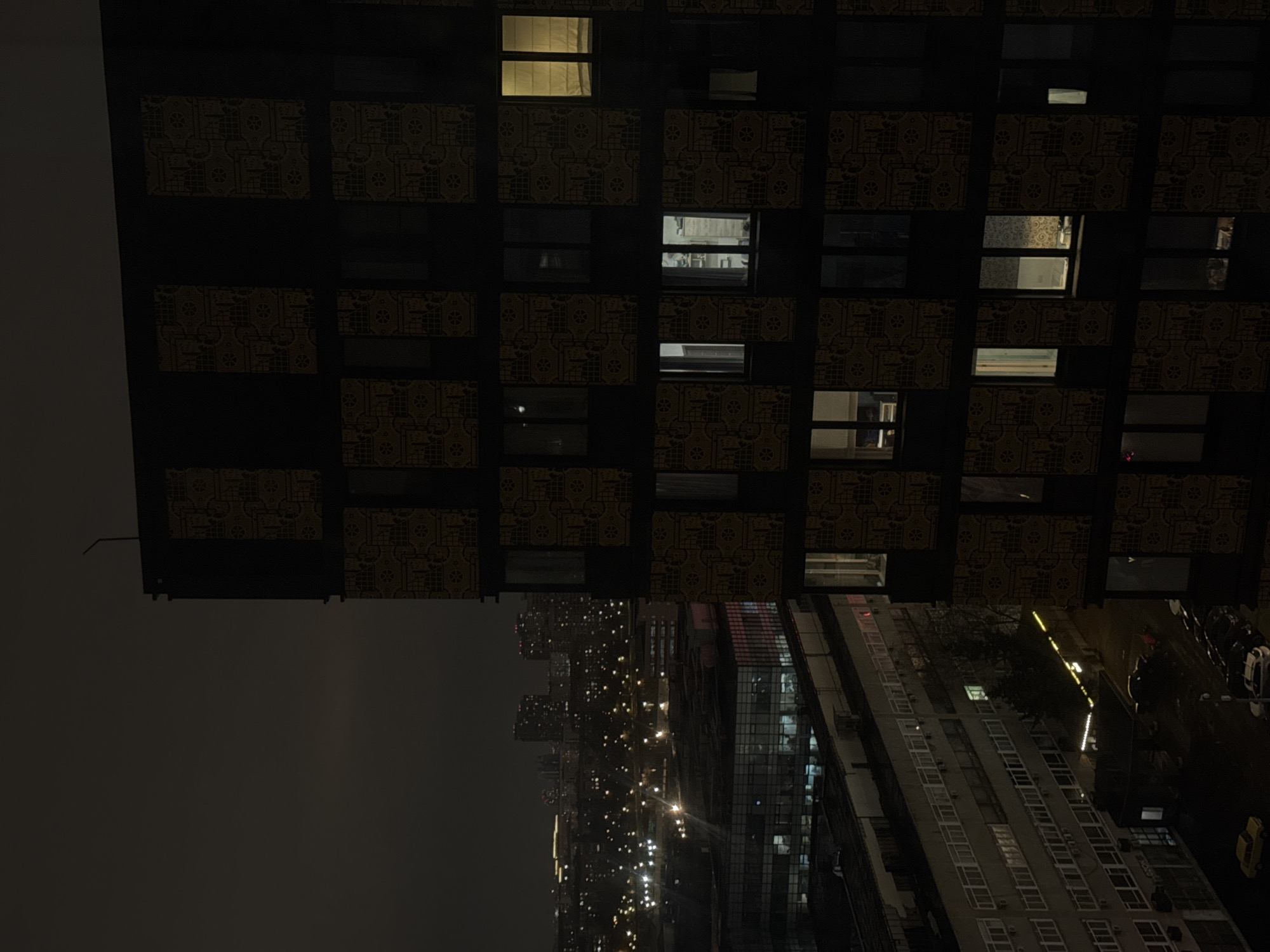Here's a description of the image:
The image shows a night view from a high vantage point in Moscow, Russia. In the foreground, a modern building with a grid-like facade is partially visible. This building is decorated with patterns, and some windows are illuminated. Below, a cityscape unfolds, with numerous buildings and streetlights visible. The overall lighting is dim, with artificial lights illuminating the scene. The buildings in the distance are a mix of residential and commercial structures, and cars can be seen on the street. The sky is dark and overcast.