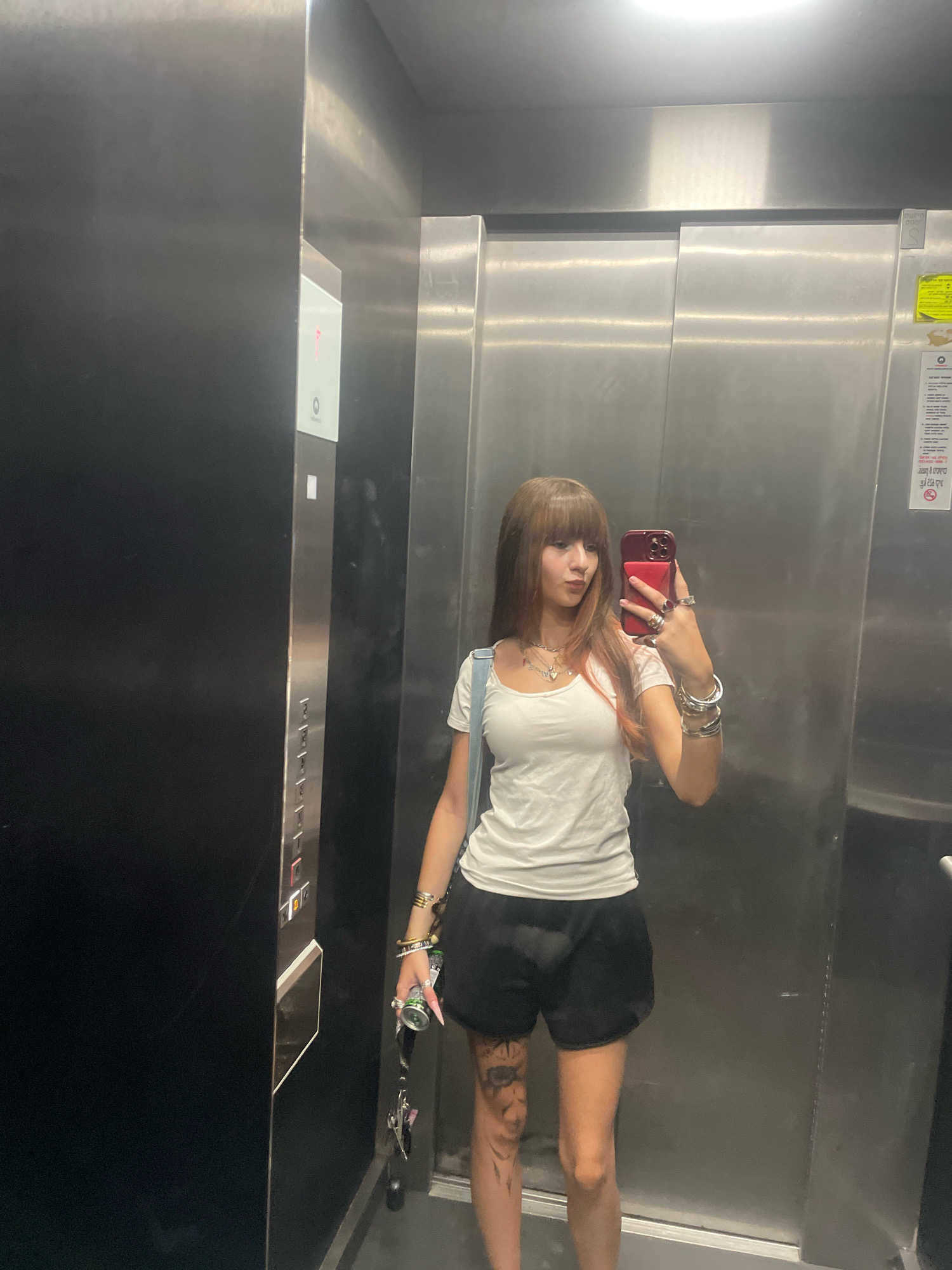A young woman with long, brown hair and bangs is taking a mirror selfie in an elevator. She is wearing a white t-shirt, black shorts, and several bracelets and rings. She is holding a red smartphone in her right hand. The elevator has stainless steel walls and a panel of buttons on the left side. The elevator appears to be located in Raanana, Israel, as indicated by the text on a yellow sticker on the right wall. The sticker, written in Hebrew, appears to be a safety notice, and the phone number displayed on it is consistent with Israeli phone numbers.
