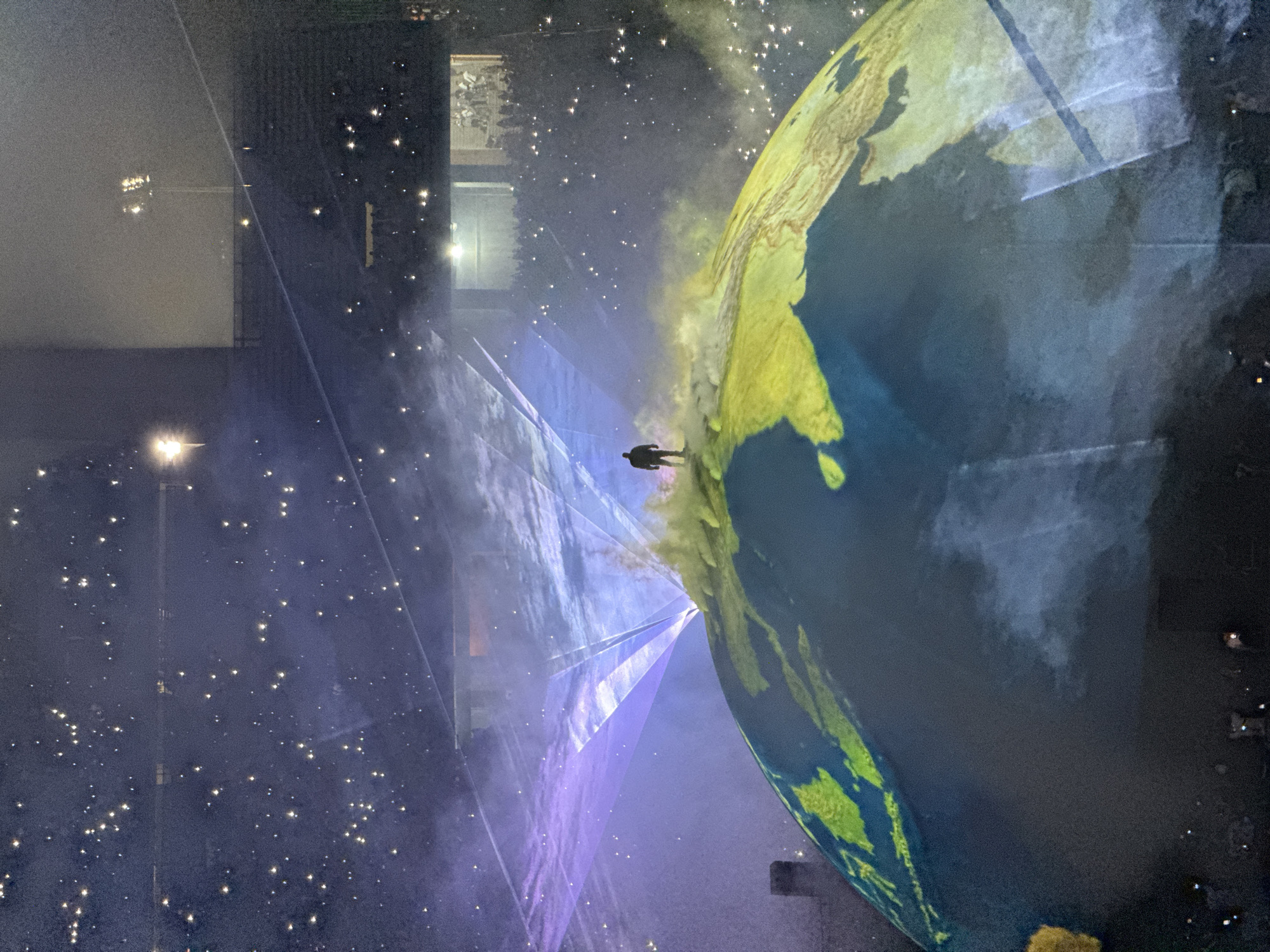 The image depicts a concert or performance setting. A person stands atop a large, globe-like structure, which appears to be a projection or inflatable displaying a map of Earth, particularly showing the Indian subcontinent. The background is filled with a starry effect, possibly created by lighting or projections. Laser beams of purple light converge towards the person and the globe. Seated audience members are visible in the upper left, suggesting an indoor arena or stadium setting. The event is taking place in Başakşehir, Turkey.