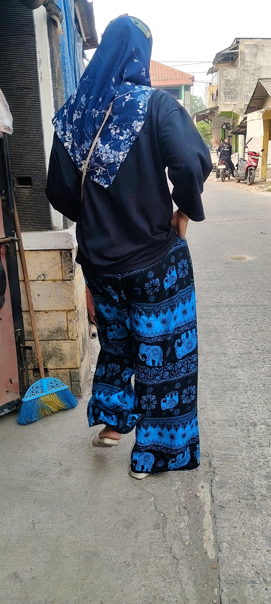 An outdoor, medium shot from a low angle shows the back of a person wearing a blue hijab with a floral pattern, a black long-sleeved shirt, and loose-fitting black pants with a vibrant blue elephant and floral pattern, walking on a paved street in Mekarmukti, Indonesia. The person's right hand is in their pocket. To the left, a broom with a blue handle and yellow and blue bristles rests against a textured wall of light-colored bricks and a dark grey ribbed surface. In the background, the street curves slightly to the right, with buildings and trees visible. A person on a motorcycle is parked on the right side of the street, with a red scooter parked next to it. The sky is overcast.