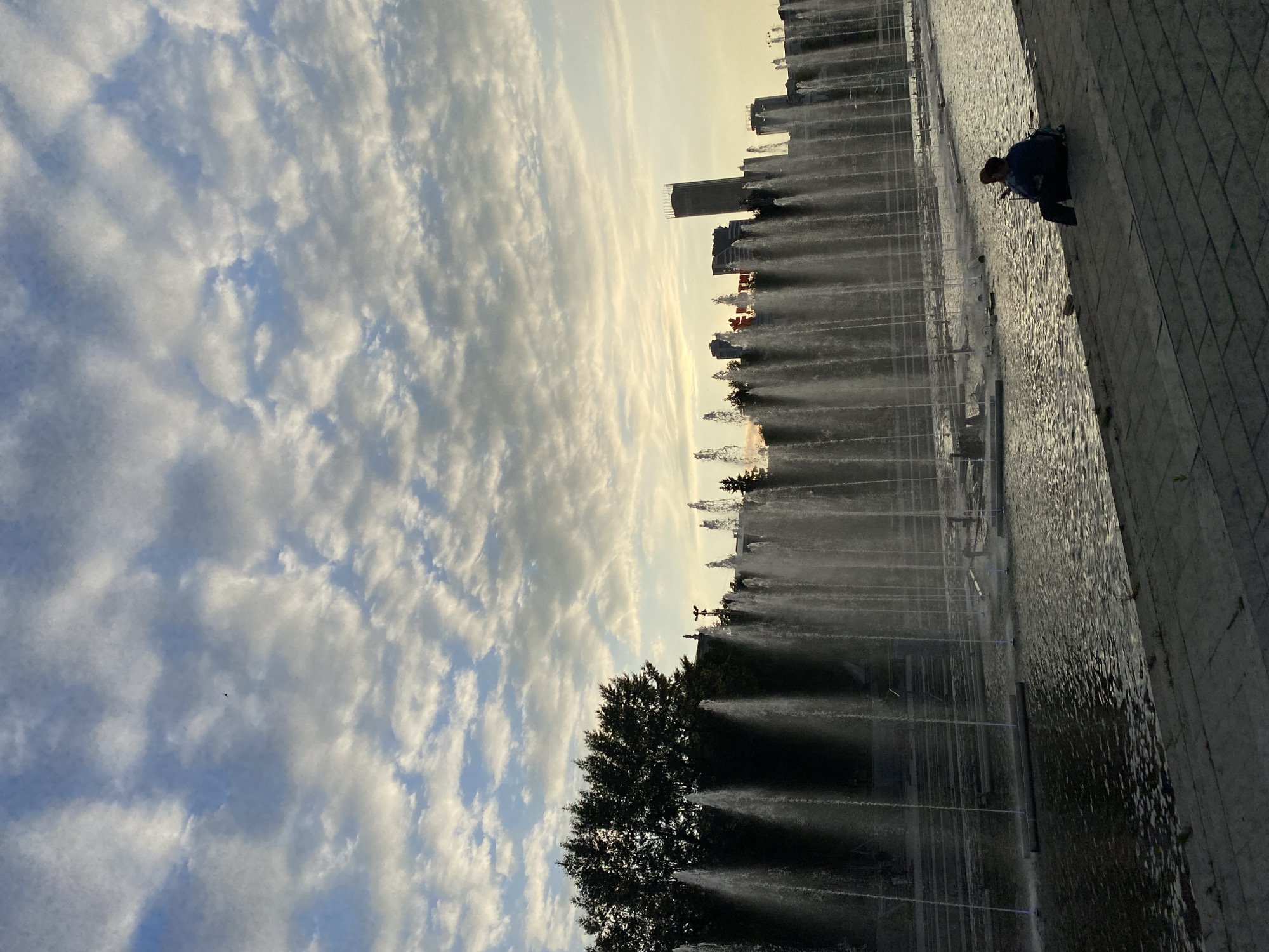The image depicts a long, multi-tiered fountain in Yekaterinburg, Russia, with water jets arcing upwards against a cloudy sky. Two people are seated on the paved embankment in the foreground, facing the fountain. In the background, trees and modern buildings, including a tall, cylindrical tower, are visible. The scene appears to be set during the golden hour, with soft, warm light illuminating the sky and reflecting on the water. The fountain stretches across a significant portion of the frame, creating a sense of scale and grandeur. The people are silhouetted against the backdrop of the fountain and the city skyline, adding a human element to the expansive landscape.