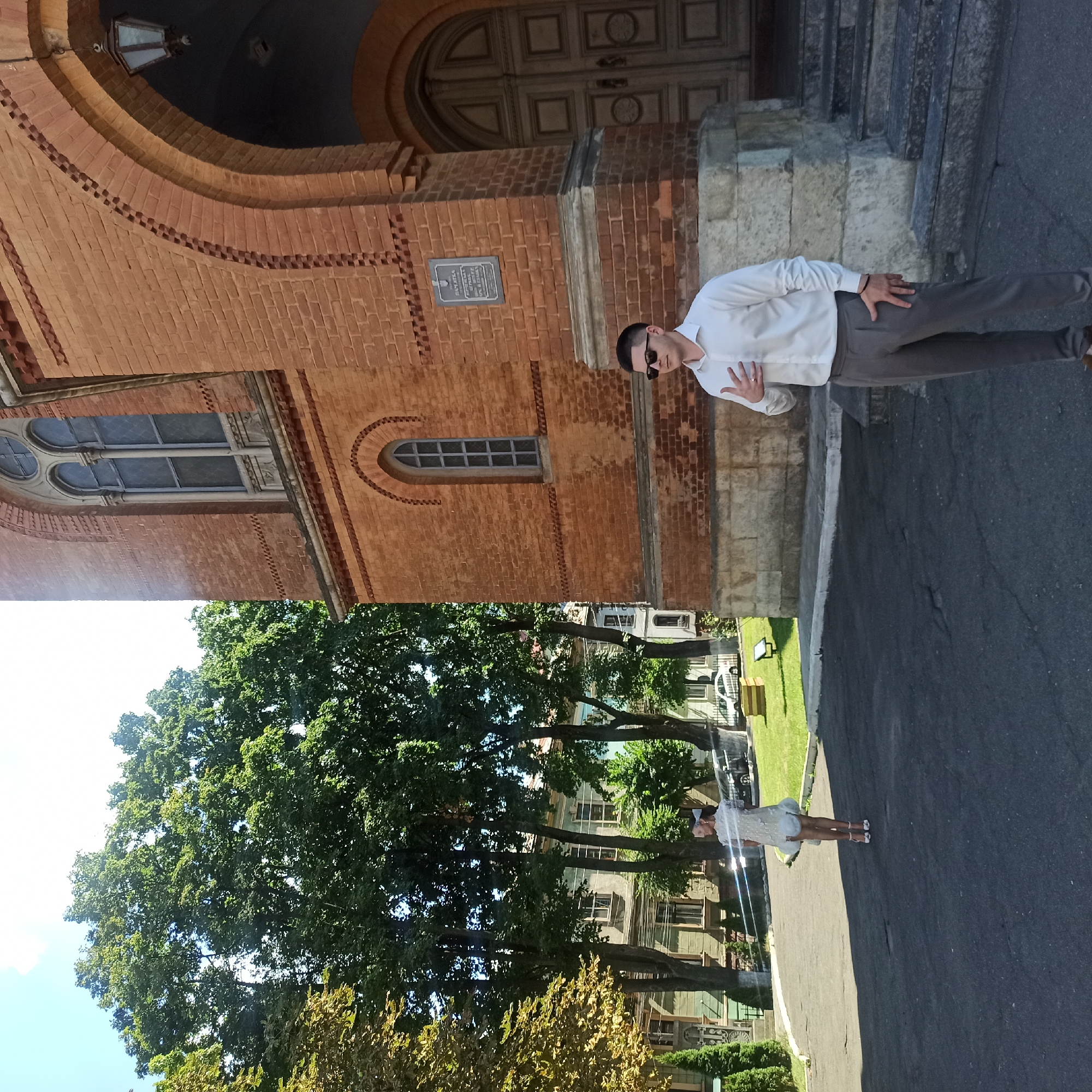 This image depicts a sunny day outside a brick building in Chernivtsi, Ukraine. In the foreground, a man in a white collared shirt and gray pants stands facing the viewer, with his left hand on his chest and his right hand near his hip. He wears sunglasses. To the left and further back, a woman in a white dress and white headband stands facing away from the viewer, her arms outstretched. She is wearing high heels. Behind the people, a large brick building with arched doorways and windows dominates the right side of the frame. Trees and other buildings are visible in the background. The ground is paved asphalt.