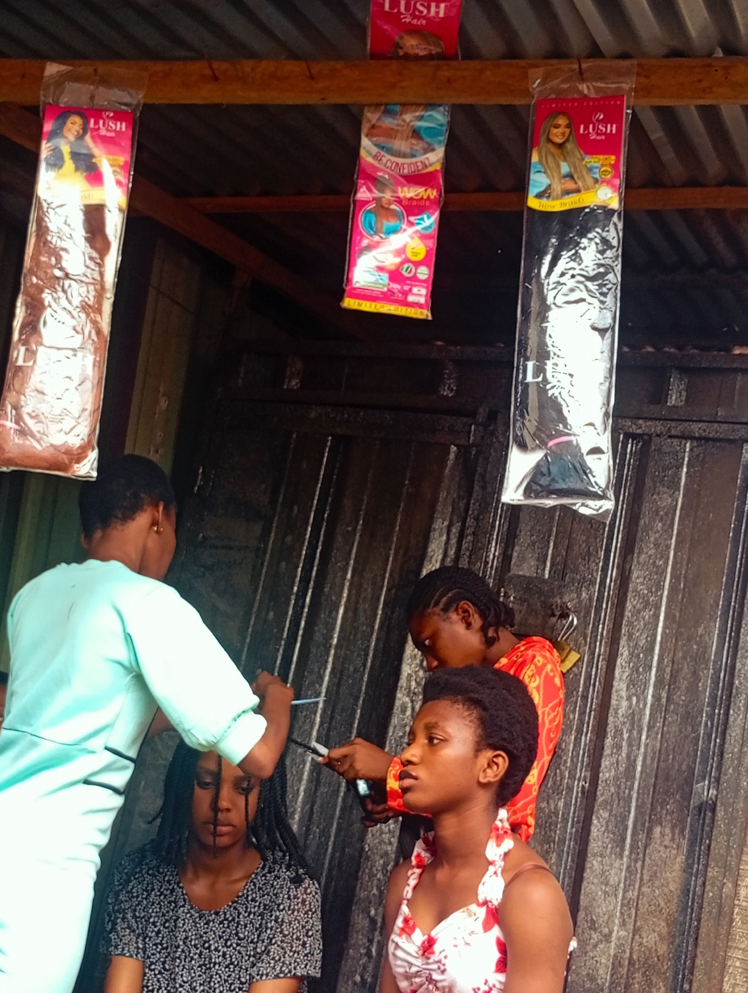 The image shows three young women in what appears to be an outdoor market stall in Nkpor, Nigeria. The stall has a corrugated metal roof and walls, with wooden beams supporting the roof. Hanging from the beams are packages of synthetic hair extensions, prominently displaying the brand "LUSH Hair". One package shows a woman with long, dark braided hair and the text "WOW Braids" and "BE CONFIDENT". Another package shows a woman with long, blonde, straightened hair, and the text "Wow Braids".
In the foreground, one woman, wearing a light green top, is in the process of braiding or styling the hair of another woman seated in front of her. She holds a pair of scissors in her right hand, suggesting she might be trimming the hair. The woman being styled has dark braided hair and is wearing a black and white patterned top. Her expression is neutral.
Behind them, a third woman, wearing a vibrant orange and red patterned top, is also assisting or observing the hair styling. She is looking down at the hair being worked on.
The overall scene suggests a hair salon or braiding stall operating in a casual, open-air environment. The lighting indicates it is daytime. The focus on hair extensions and the activity of hair braiding are central to the image.