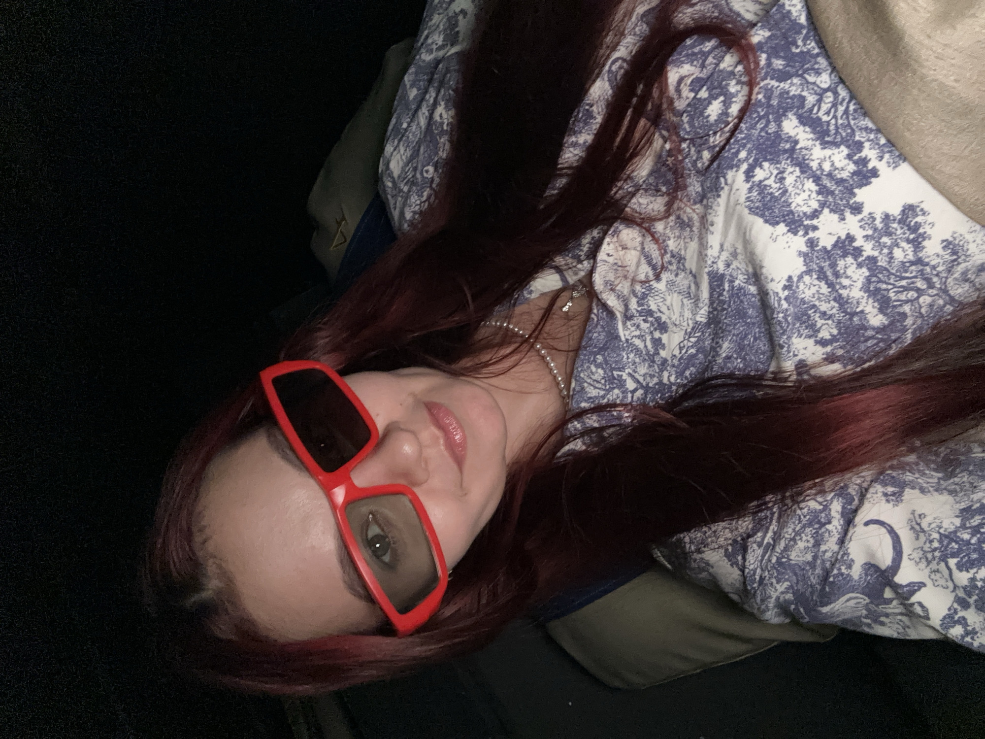 A woman with long, dark red hair, wearing red-rimmed 3D glasses and a blue and white patterned shirt, poses for a selfie. She is wearing a pearl necklace with a small diamond pendant. The background is dark and appears to be indoors, possibly in a dimly lit room. There are no visible landmarks or contextual clues to indicate the location is Khan Daun Penh, Cambodia.