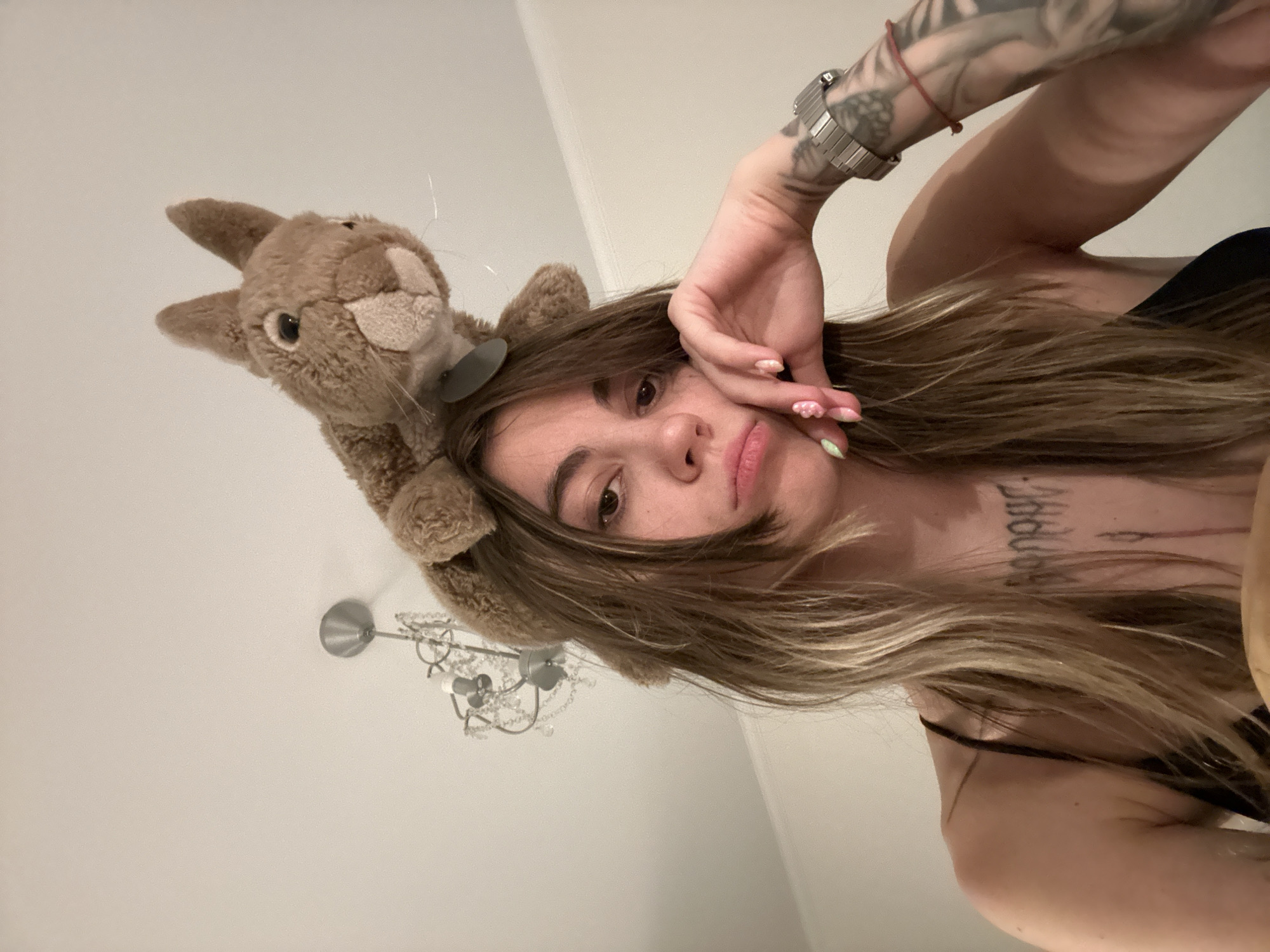 This media file features a young woman taking a selfie in what appears to be a bedroom. She is lying down, with her head tilted upwards towards the camera. On her head, she is wearing a plush brown bunny headband. Her right hand is raised, with her fingers lightly touching her face and hair. Her left arm is also visible, extended upwards, showing tattoos on her arm and a silver watch on her wrist. A delicate red bracelet is also visible on her left wrist. She has long, wavy, light brown hair that falls around her face and shoulders. Her lips are slightly parted, and she is looking directly at the camera with a neutral expression.
In the background, a silver chandelier with crystal accents is visible on the ceiling, suggesting an indoor setting, possibly at night or in a room with limited natural light. The walls are a plain, light color. The overall mood is casual and intimate. There is no visible text in the image. The location context is Moscow, Russia.