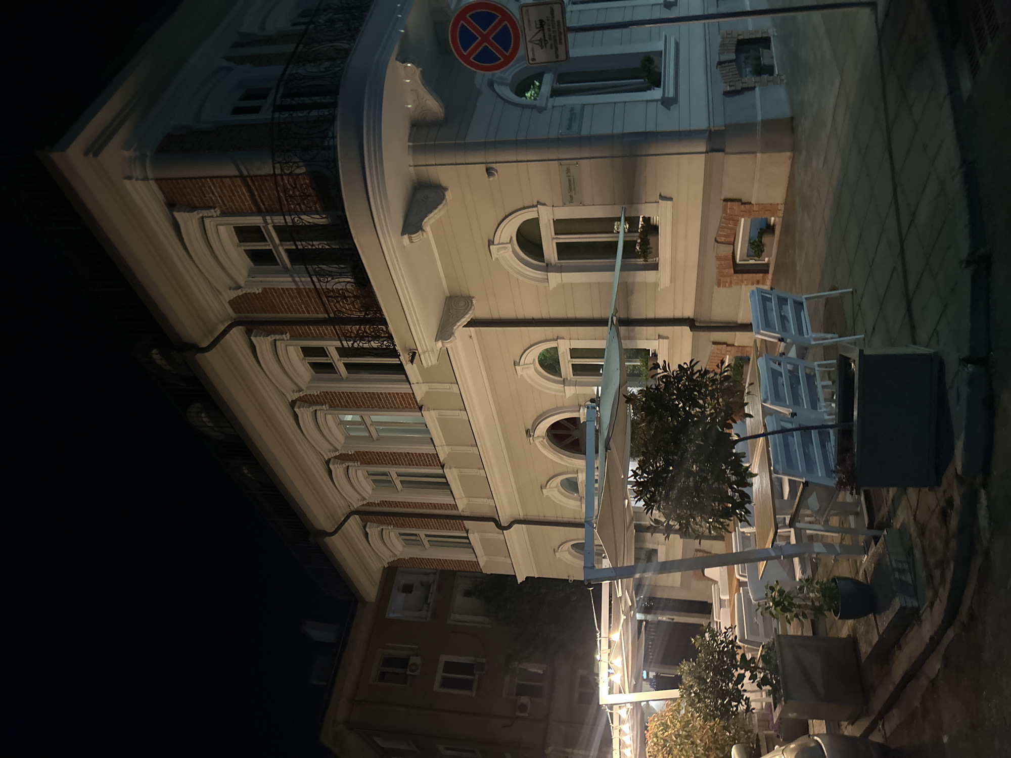 The image depicts a street scene in Burgas, Bulgaria at night. A multi-story building with ornate architectural details, including arched windows, brick accents, and decorative molding, dominates the view. A red and blue no-parking sign is visible on the corner of the building. Along the sidewalk in front of the building, there is an outdoor seating area with white chairs and tables under awnings. Small trees in pots are placed around the seating area. The right side of the image shows a stone-paved sidewalk and the left shows a dark, unlit sky.