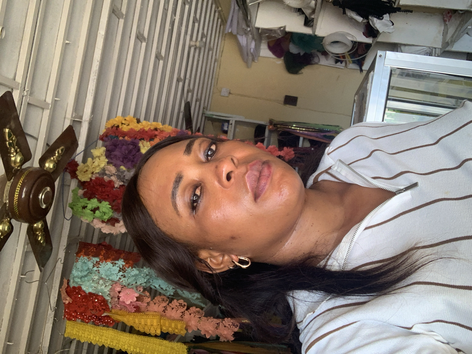 A woman with dark hair and brown eyes is pictured in Sabon Tasha, Nigeria, wearing a white and brown striped shirt. She is wearing gold earrings. There is a ceiling fan next to her, and colorful fabrics are hanging behind her. A glass display case is visible in the background.