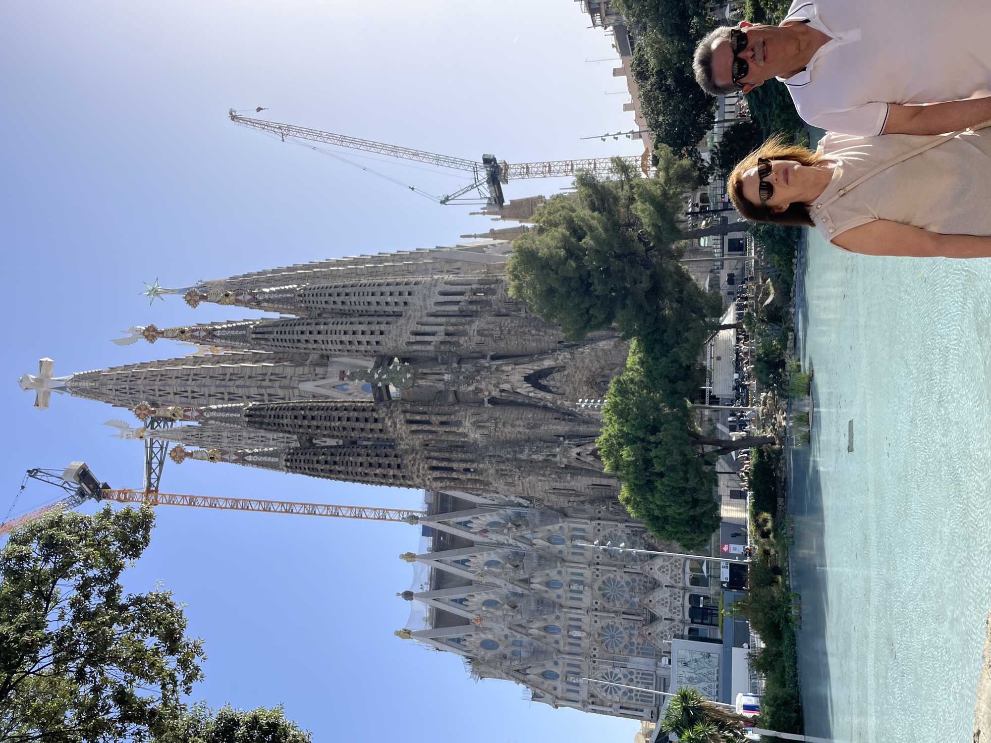 The image shows a couple standing in front of a large, ornate church with several spires, identified as the Sagrada Familia basilica in Barcelona, Spain. The basilica is under construction, with large cranes visible in the background, indicating ongoing work. In the foreground, there is a body of light blue water, possibly a reflection pool or fountain, with green trees and landscaping around it. The couple appears to be tourists, dressed casually and wearing sunglasses. The sun is shining, casting shadows and creating a bright, clear day. The background includes more trees and buildings.