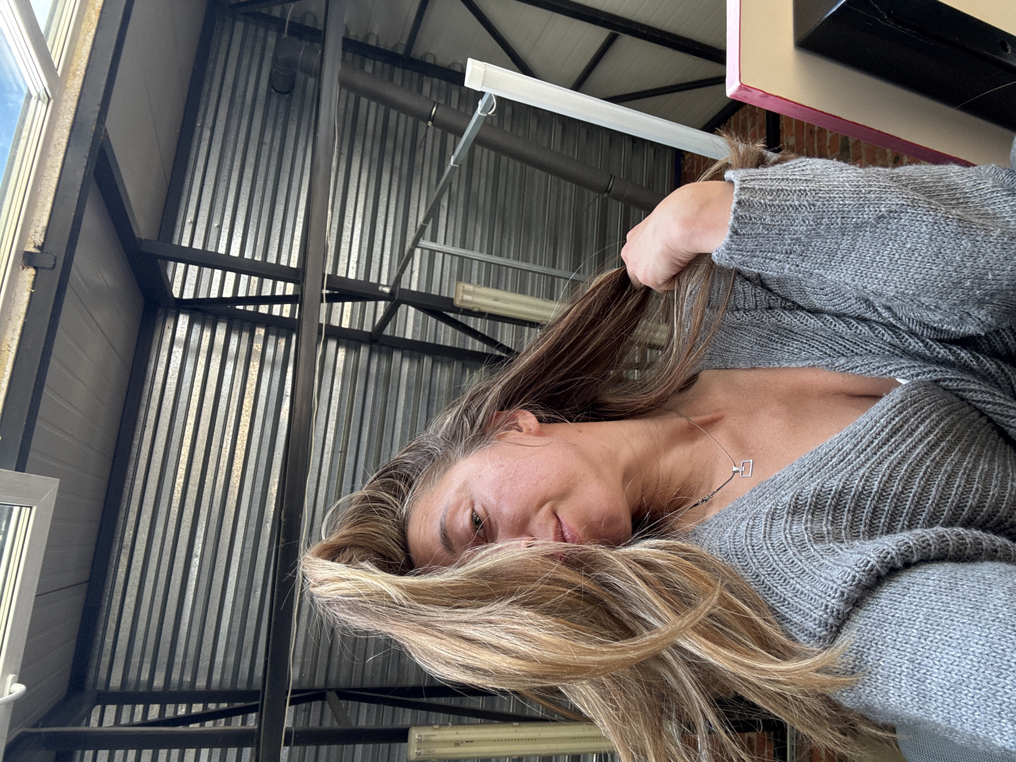 A woman with long, blonde hair is looking at the camera in a selfie. She is wearing a grey knitted sweater and a necklace with a geometric pendant. Her hair is blowing in her face, partially obscuring her left eye. The background appears to be an industrial or warehouse-like setting, with corrugated metal walls and exposed metal beams. There is a skylight visible at the top of the image, showing a glimpse of the sky. The scene is indoors, possibly a workshop or studio, with some light sources visible on the ceiling. The overall lighting is natural, coming from the skylight and possibly other unseen sources.
