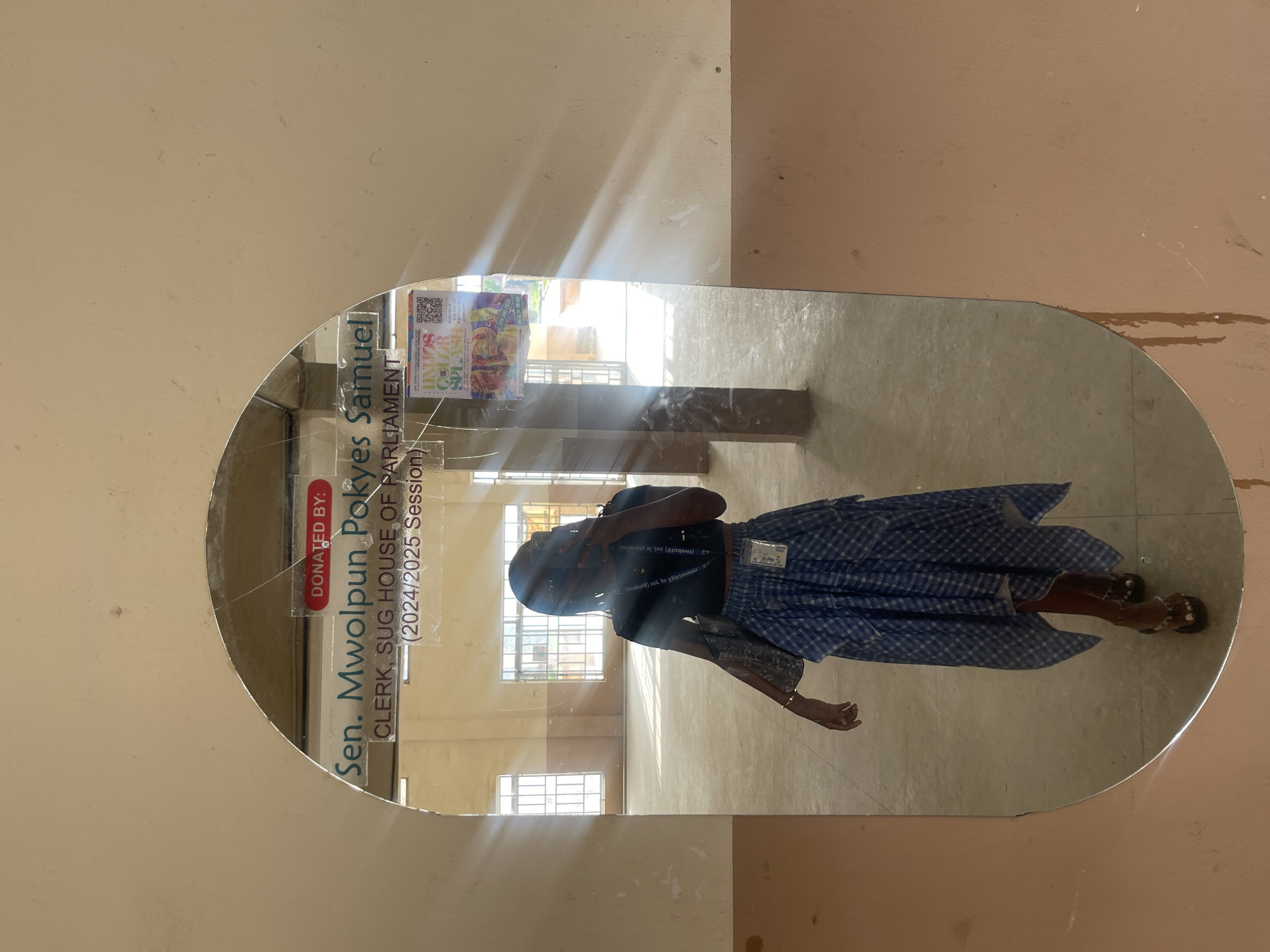 A woman stands in front of a cracked mirror in what appears to be a vacant building. The mirror reflects her from the waist down, showing her wearing a blue checkered skirt and brown sandals. Above her, a sign reads "DONATED BY: Sen. Mwolpun Pokyes Samuel CLERK, SUG HOUSE OF PARLIAMENT (2024/2025 Session)". To the right of the sign, a colorful poster is visible. Sunlight streams through windows in the background, illuminating the dusty, empty space. The walls of the building are painted in light brown and beige tones. The overall impression is one of a dimly lit, possibly abandoned or under renovation, interior. The location is identified as Rafin Par, Nigeria.