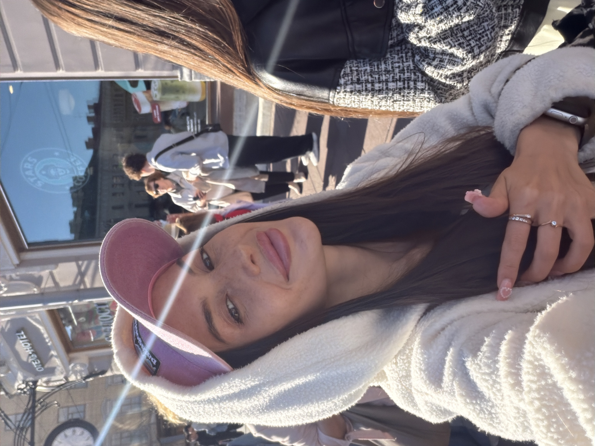 The image is a selfie taken outdoors in a city environment, likely Saint Petersburg, Russia, given the Cyrillic signage visible on a building. The foreground subject is a young woman with long dark hair, wearing a white hooded jacket and a pink baseball cap. She is looking at the camera and smiling, with her right hand resting on her chest, adorned with rings and polished fingernails. To her right, another young woman with long brown hair is partially visible, wearing a black and white tweed jacket over a black leather jacket. In the background, a cafe with a "STARS COFFEE" logo is visible through a large window, reflecting buildings and people outside. Other individuals can be seen in the background, as well as a clock on a building facade and street lamps. The lighting suggests it is daytime, with sunlight casting shadows and lens flares.