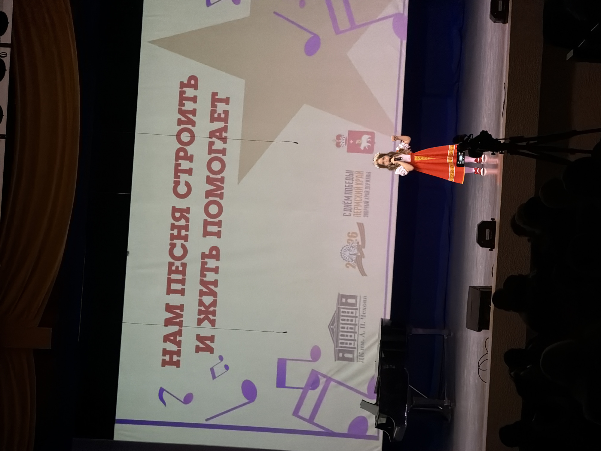 A wide, long shot shows a young female performer on a stage, captured in vertical orientation. The performer is dressed in a red, knee-length dress adorned with gold trim and white undersleeves, a white headpiece, and red shoes.
Behind her, a large screen displays the Russian text "2 НАМ ПЕСНЯ СТРОИТЬ И ЖИТЬ ПОМОГАЕТ", meaning "Songs help us build and live". Below this is the text "ДК им. А. П. Чехона", likely referring to the A.P. Chekhov Cultural Center, and "С ДНЁМ ПОБЕДЫІ ПЕРМСКИЙ КРАЙ ОПОРНЫЙ КРАЙ ДЕРЖАВЫ", meaning "Happy Victory Day! Perm Territory is the stronghold of the state."
The screen also features illustrations of musical notes, the emblem of Perm, and the number "2026" alongside a stylized image of a cannon, possibly indicating an anniversary.
In front of the performer, a video camera on a tripod captures her performance. The lower right corner of the image reveals a portion of the audience in silhouette. Stage lights and speakers are also visible. The location of this event is Perm, Russia.