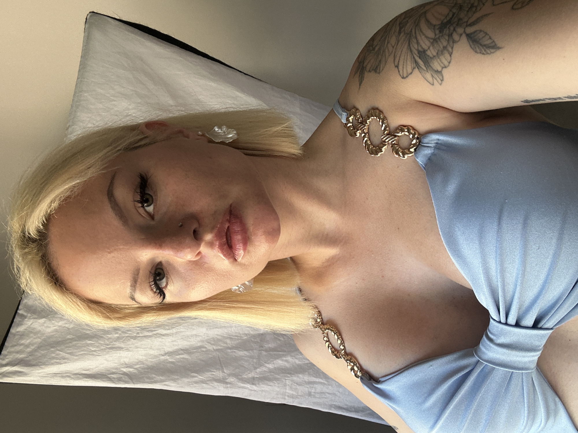 A woman with blonde hair and noticeable makeup is posing for a selfie in what appears to be a hotel room or studio in Budva, Montenegro. She is wearing a light blue, low-cut dress with gold chain detailing on the straps and a large bow in the center. The background consists of a white fabric backdrop and a light-colored wall. The woman has a pierced nose and a large tattoo on her left arm. She is looking directly at the camera with full lips and long eyelashes.
