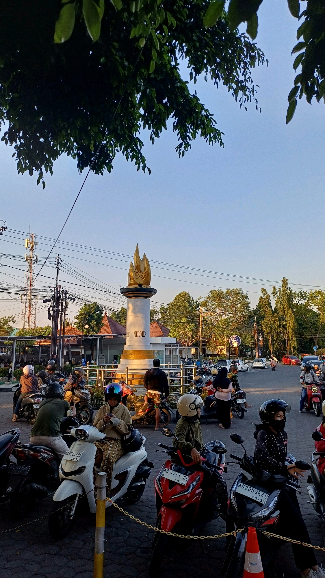 The image depicts the Tugu Merdeka (Freedom Monument) in Klaten, Indonesia. The monument, characterized by its golden flame atop a white cylindrical base with gold accents, stands prominently in the center of a traffic circle. Surrounding the monument are numerous motorcycles, with riders seated or standing, suggesting a gathering or waiting period. Several people are visible, some wearing helmets, engaged in various activities like looking at their phones or conversing. The environment features utility poles with a complex network of wires, trees, and buildings in the background, including a cell tower on the left. The scene is bathed in the warm light of what appears to be late afternoon or early morning, casting long shadows. The ground is paved, and chain barriers are visible around the monument. Cars are also present in the background, indicating a moderately busy urban intersection.