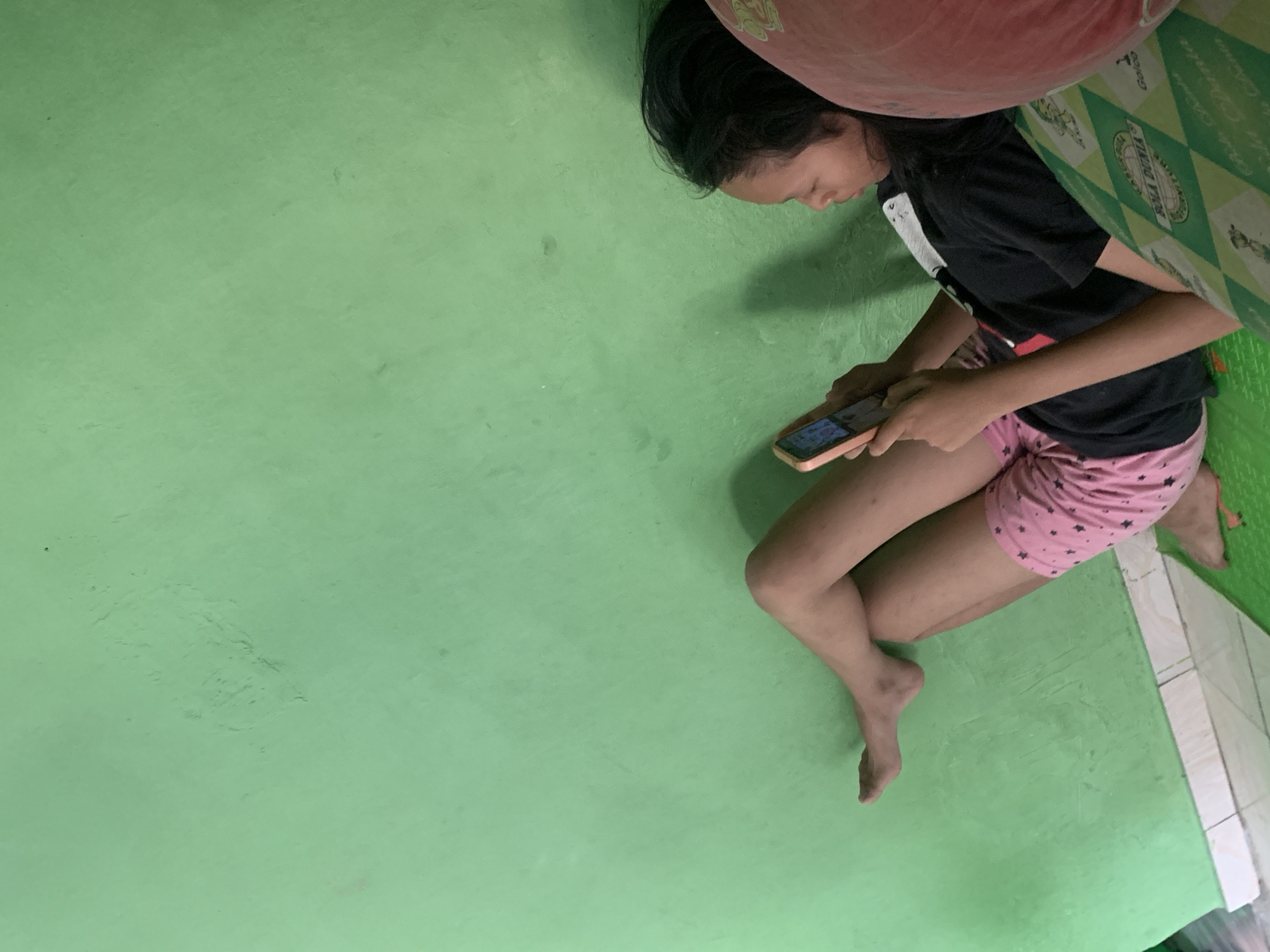 A young Indonesian girl is depicted lying down and using a smartphone. She is positioned on her left side, with her legs crossed and her right hand holding the phone. She wears a black t-shirt and pink patterned shorts. The background is a plain green wall. The scene appears to be in a residential setting, possibly a bedroom, and the lighting suggests it is daytime. Based on the available information, this scene is likely located in Palembang, Indonesia.