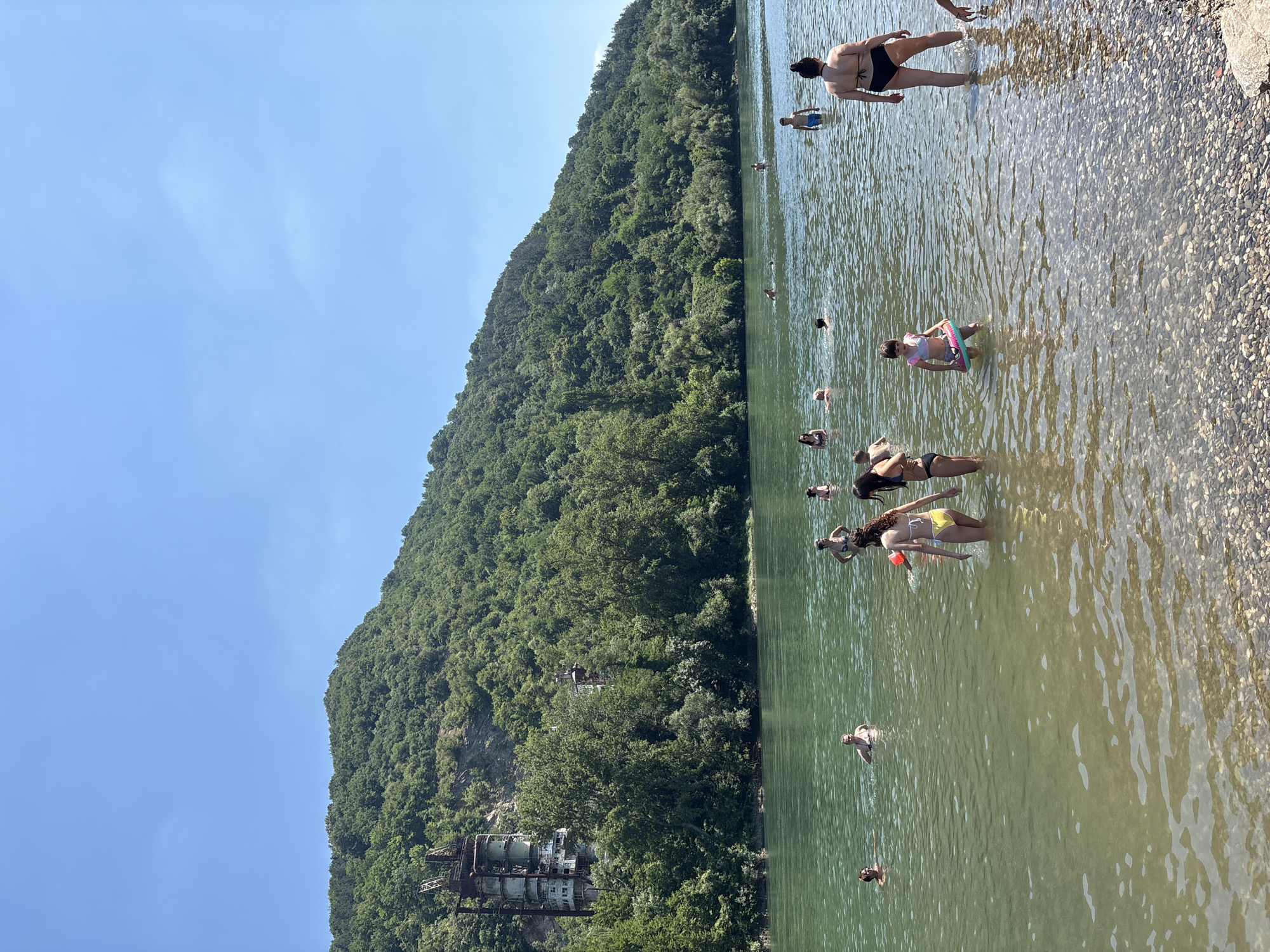 A wide shot captures a clear, sunny day at a river in Zvornik, Bosnia and Herzegovina, with a steep, tree-covered hill dominating the background. The hill features an abandoned, multi-story concrete industrial structure near its base on the left side, with visible metal scaffolding and debris. The river itself is populated with numerous people swimming and wading in the greenish water, some in swimsuits and others with inflatable toys. The riverbed is visible in the foreground due to shallow water and is covered in pebbles. The sky is a bright, cloudless blue.