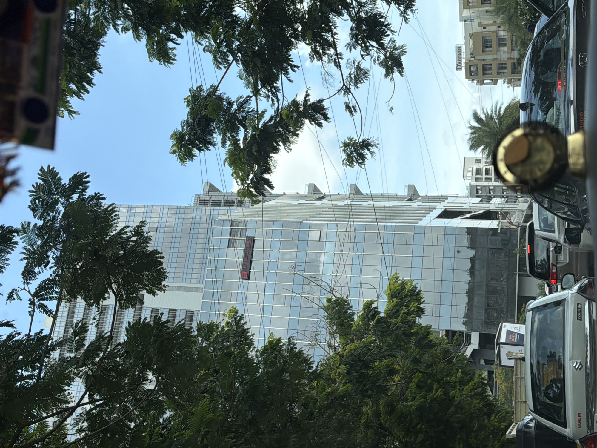 The image captures a street scene in Hyderabad, India, dominated by a modern glass-fronted skyscraper. The building features multiple stories with reflective windows that mirror the blue sky and surrounding trees. A red sign with white text, possibly a company name or advertisement, is visible on the building's facade. Lush green trees, with dense foliage, frame the scene from the left and top, obscuring parts of the sky and building. Power lines crisscross the sky, a common sight in urban Indian environments. In the foreground, several cars are seen on a road, indicating traffic. One car in the immediate foreground is a silver hatchback with a visible logo that reads "MARUTI SUZUKI." Another car, a darker SUV, is partially visible behind it. The overall atmosphere suggests a busy urban environment under a bright sky, with a juxtaposition of natural elements and modern architecture.