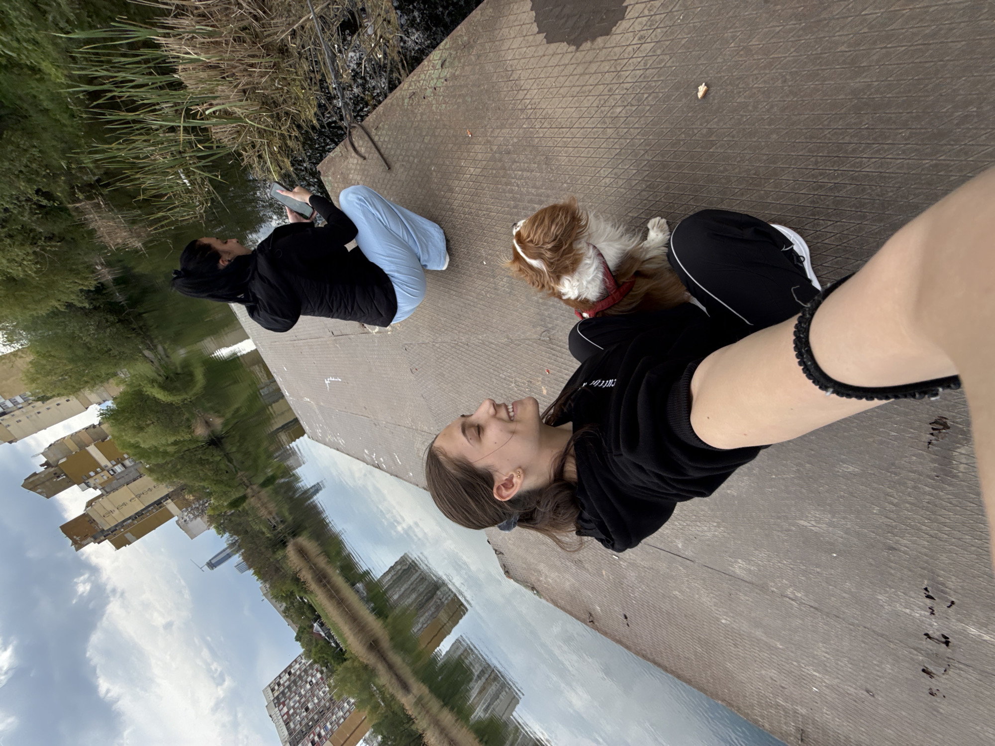 This image captures two individuals and a dog relaxing on a textured concrete platform beside a calm body of water, likely a canal or river, in Sofia, Bulgaria. The perspective suggests a selfie.
In the foreground, a young woman with brown hair, wearing a black hoodie and black athletic pants, smiles towards the camera. She wears earrings and a black beaded bracelet. Beside her, a small, brown and white dog, wearing a red harness, lies calmly on the platform. Further back, another person sits on the platform, mostly facing away from the camera, wearing a black top and light blue pants, engaged with their smartphone.
The water clearly reflects a partly cloudy sky and distant urban buildings, including multi-story structures and one with a crane, indicating an urban or suburban environment. The water's edge is lined with green reeds and grass, with trees visible beyond. The platform itself appears worn, with a diamond-grid pattern, and a small piece of bread or food is visible on its surface. The scene takes place during the daytime under what appears to be an overcast sky, creating soft, even lighting. The overall mood is relaxed and peaceful, highlighting a casual outing.
