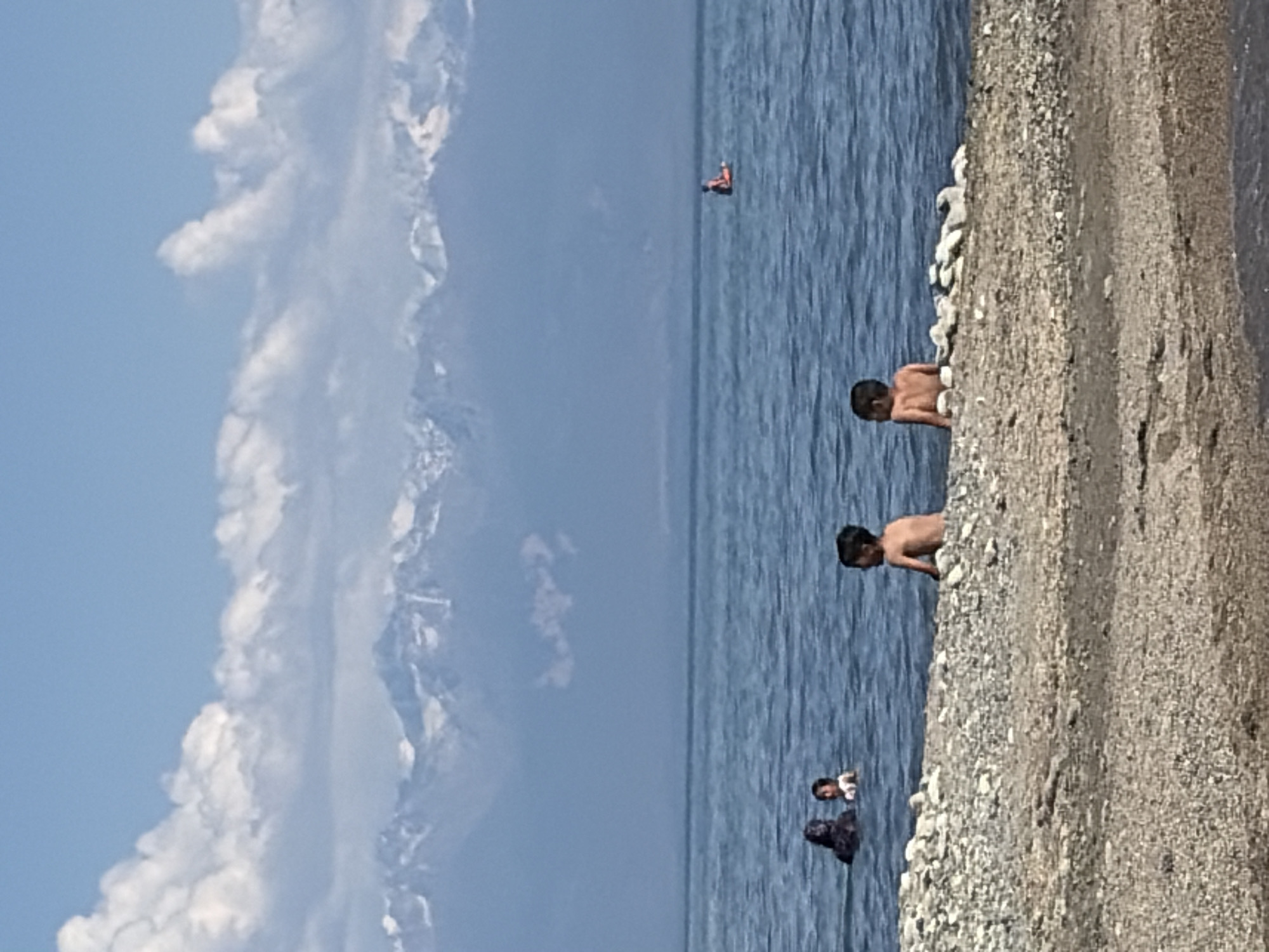 The image depicts a scene at a beach with clear blue water and a gravelly shore. The sky is a bright blue with fluffy white clouds. In the water, there are several people. Two young boys are visible near the shore, one sitting on the gravel and looking out at the water, and another wading in the shallow water. Further out in the water, a person is sitting on a float or small raft. In the background, distant mountains can be seen, their peaks covered in snow or white rock. The water is calm and appears to be a lake or a sheltered bay. The overall mood is relaxed and summery, suggesting a pleasant day at the beach. The time of day appears to be daytime, given the bright sunlight and clear skies. There is no visible text in the image.