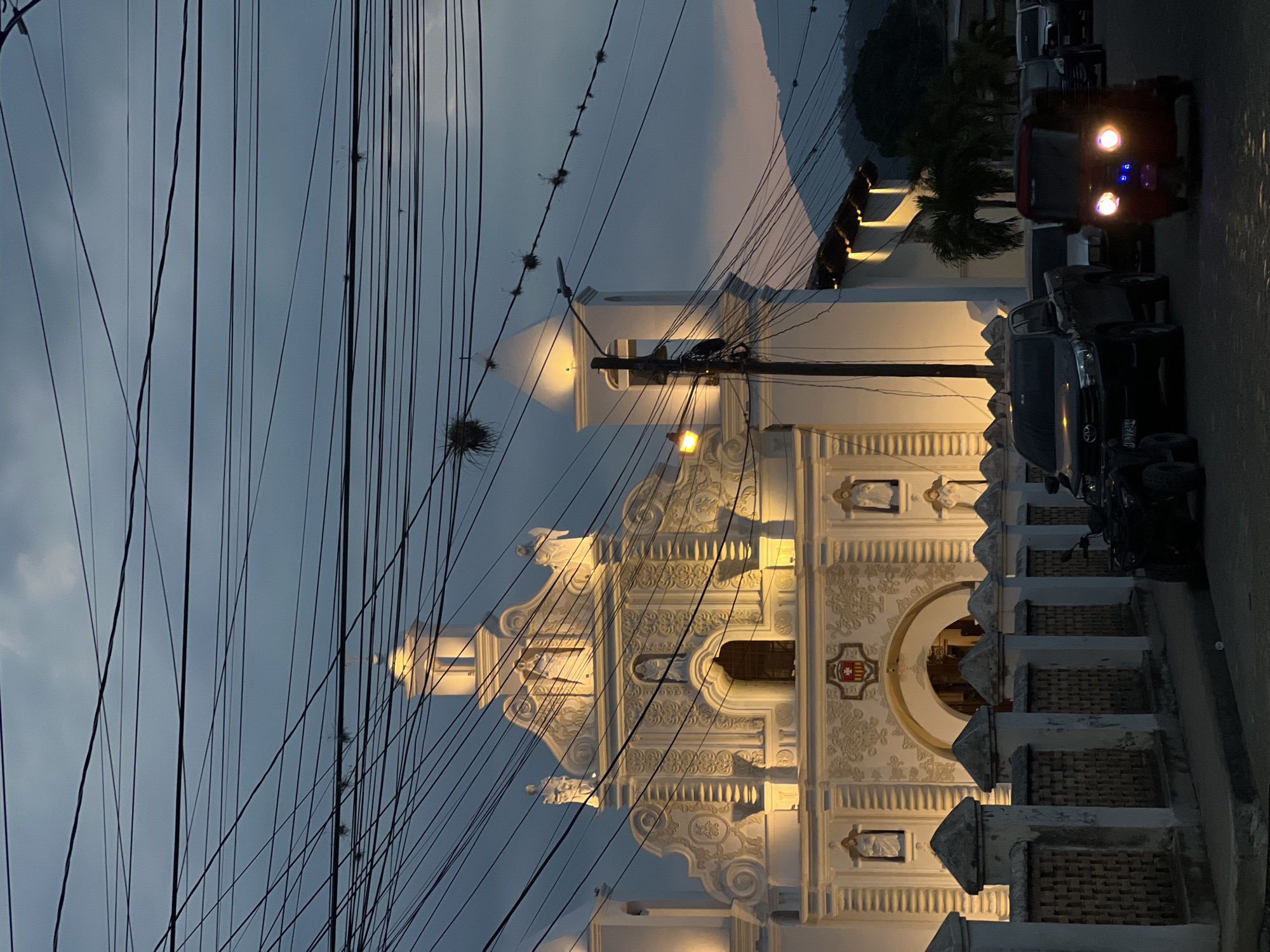 The image captures the Iglesia de San Sebastian in Gracias, Honduras. The white, ornate facade of the church is illuminated, standing out against the dusk sky. Power lines heavily crisscross the view, partially obscuring the church's architecture. To the right, a row of parked vehicles lines the street. A red jeep with bright headlights is visible. The environment suggests a quiet evening in the city.