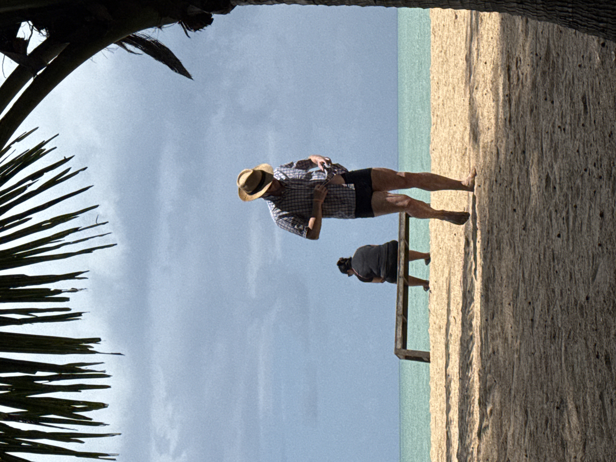 A man wearing a straw hat, checkered shirt, and dark shorts walks on a sandy beach in Ban Khuk Khak, Thailand. He is adjusting the front of his shirt. Further back, a person wearing a dark shirt and shorts sits on a wooden bench, facing away from the viewer, looking out at the ocean. The scene is framed by palm fronds at the top and a palm tree trunk on the right. The sky is overcast and the sea is a calm turquoise. The sand is light brown with scattered dark specks. Shadows from the palm fronds cast stripes across the sand in the foreground.