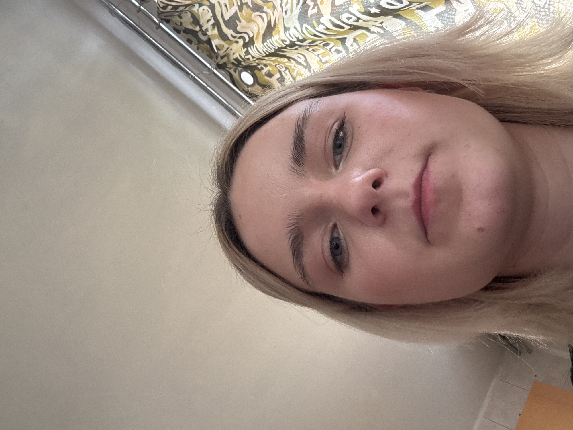This is a close-up, tilted portrait of a young woman with blonde hair, fair skin, and blue eyes, looking directly at the camera. Her face, framed by her light hair, occupies a significant portion of the image, angled from the lower right towards the upper left. She has natural light brown eyebrows, and a small mole or blemish is visible on her right cheek (viewer's left). Her lips are slightly parted, and her expression is neutral.
The background is varied. In the upper left and central areas, there is a patterned fabric, possibly a curtain or tapestry, featuring abstract designs in shades of yellow, green, and brown. Indistinct text appears to be integrated into this pattern, with one visible section vaguely suggesting "Wonder." A metallic bar or railing runs horizontally along the upper edge, partially hidden by the fabric. The lower left and central background is a plain, light-colored surface, possibly a wall or ceiling. In the bottom right corner, a small area of light-colored tiled flooring with darker grout lines is visible, along with a piece of light brown furniture.
The lighting is soft and appears to be indoor, illuminating her face evenly without harsh shadows. The image captures a personal moment, likely a selfie or a closely taken portrait within an interior setting in Saint Petersburg, Russia.