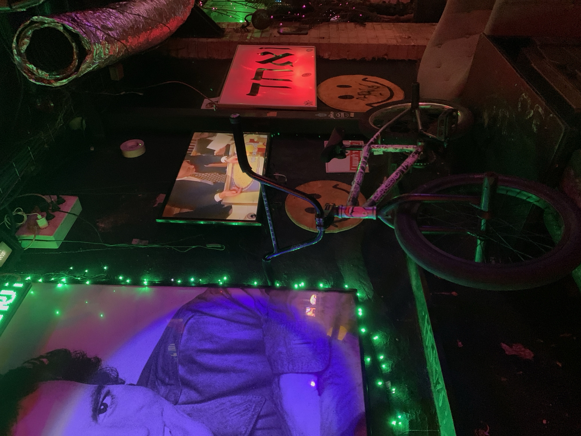 This image captures an indoor scene, possibly a bar or a venue, in Tel Aviv, Israel. The ambiance is dark, illuminated by vibrant green and red neon lights, and the glow from a television screen. Dominating the left side of the frame is a large, black-and-white portrait of a man, bathed in purple and green light. In the foreground, a BMX bicycle is positioned vertically, leaning against a surface. The bicycle is adorned with stickers, including smiley faces. Behind the bicycle, a television screen displays a scene with people in formal attire, and a red illuminated sign with Hebrew text hangs on the wall. Other elements in the scene include exposed pipes, brickwork, and a portion of upholstered seating. The overall impression is one of an eclectic, dimly lit, and potentially artistic or counter-cultural space.