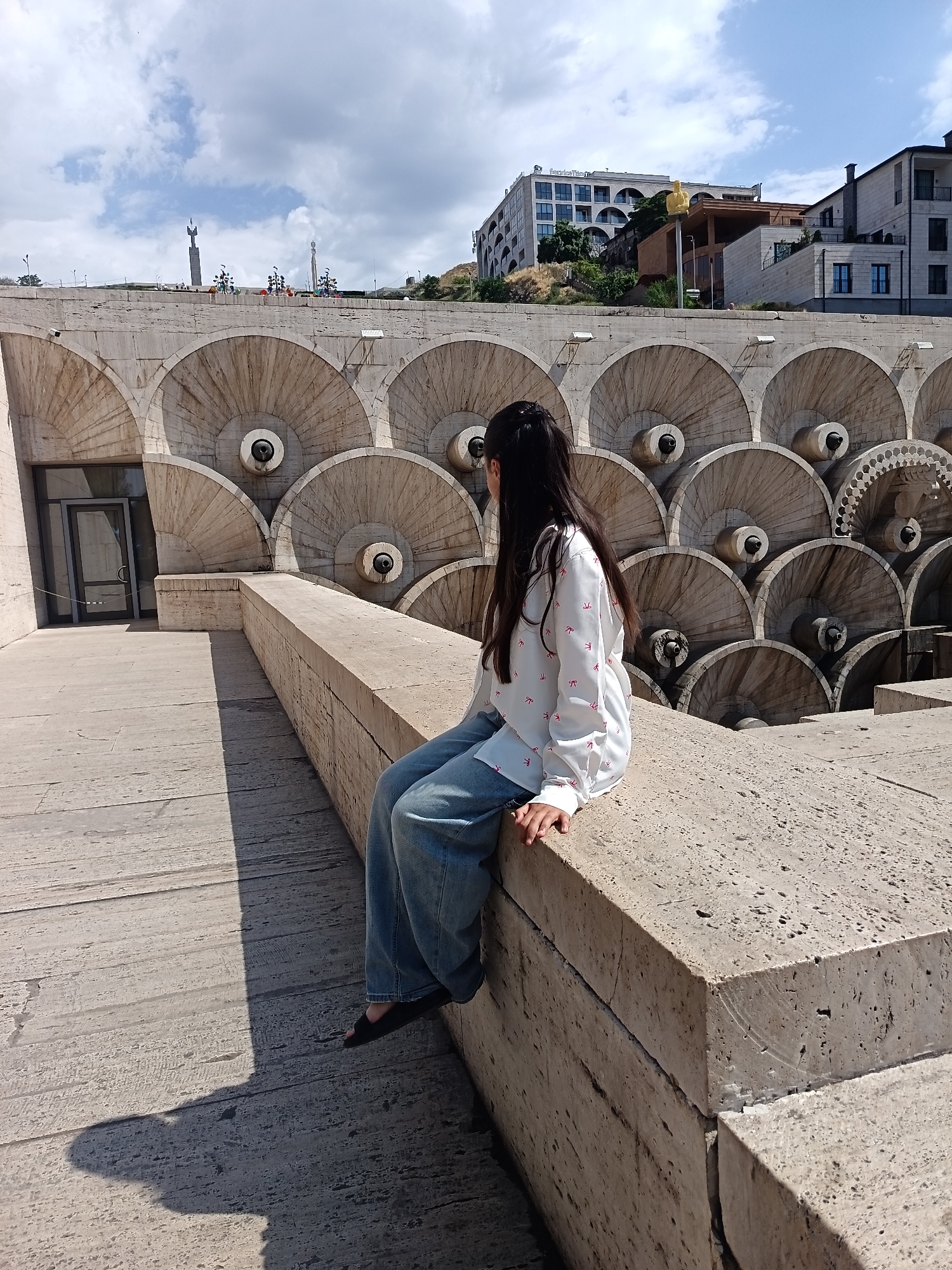 A young woman with long dark hair, seen from the side, sits on a concrete ledge. She is wearing a white long-sleeved shirt with small red bow-shaped patterns and loose-fitting blue jeans. Her feet are in dark open-toed shoes. The background features a large, tiered concrete structure with numerous semicircular indentations, some of which have dark spouts. In the distance, several buildings and monuments are visible against a cloudy blue sky. The foreground shows a concrete pathway with a distinct shadow cast by the woman.