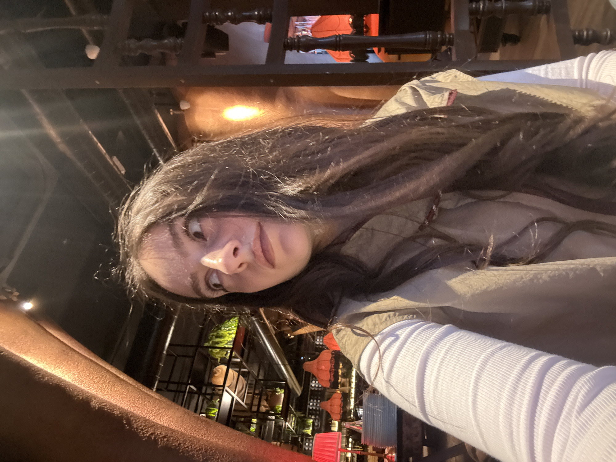 A woman is facing the camera in Yerevan, Armenia. The scene appears to be indoors, possibly a restaurant or bar. The woman has long brown hair, a light complexion, and is wearing a white long-sleeved shirt and a tan jacket. She is slightly angled, with her face prominent in the frame. The background features architectural elements of the establishment, including dark wooden beams, lighting fixtures with orange and red shades, and plants. There are other decorations that suggest a somewhat eclectic or themed design style. The overall lighting is warm, with a mix of natural and artificial light sources contributing to the ambiance.