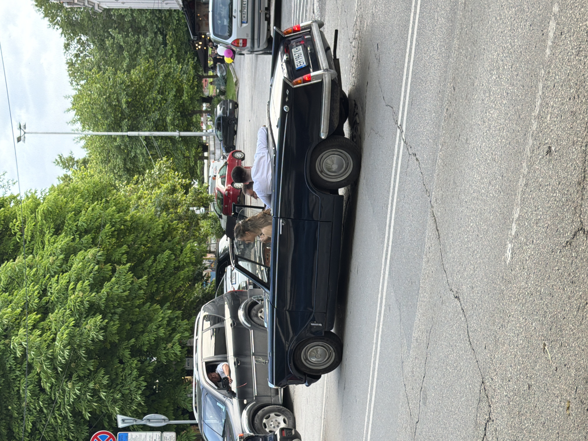 The image depicts a street scene in Pleven, Bulgaria. A dark blue vintage convertible car without a roof is parked on the side of a street with double white lines. Two individuals are visible in the car, a man in a white shirt and a woman with light hair. The car's license plate is partially visible. Other vehicles are parked ahead of and behind the convertible, including a silver SUV, a dark SUV, and a red car in the distance. Trees line the side of the street. A tall street light is visible near the trees. A street sign is also seen near the curb. The road surface shows signs of wear and cracking.