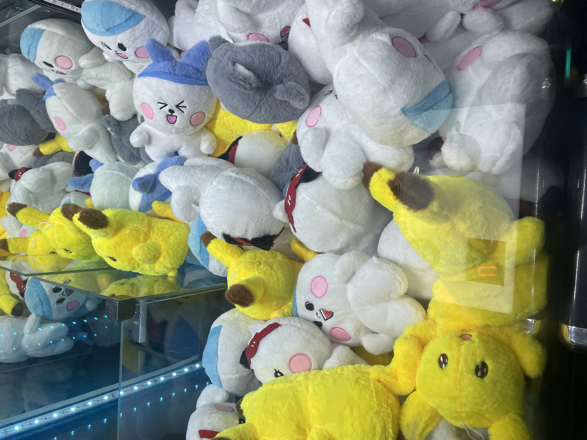 The image shows a close-up, eye-level view of a claw machine filled with various stuffed toys. The toys are densely packed and appear to be primarily white, yellow, and some gray. Dominant among the stuffed animals are yellow Pikachu-like characters with brown ears and white characters with pink cheeks and various colored accessories like blue headbands or red bandanas. Some of the white characters resemble rabbits or mice with closed eyes and simple embroidered facial features. A few gray, round stuffed animals are also visible. The claw machine itself is visible at the bottom, with a metallic edge. The background is blurred but suggests the interior of a game arcade, with a blue LED light strip running vertically on the left side of the frame. The lighting is artificial and casts some reflections on the glass of the machine. There are no people or discernible actions taking place within the frame, other than the presence of the toys themselves. The scene is static and appears to be an indoor environment.