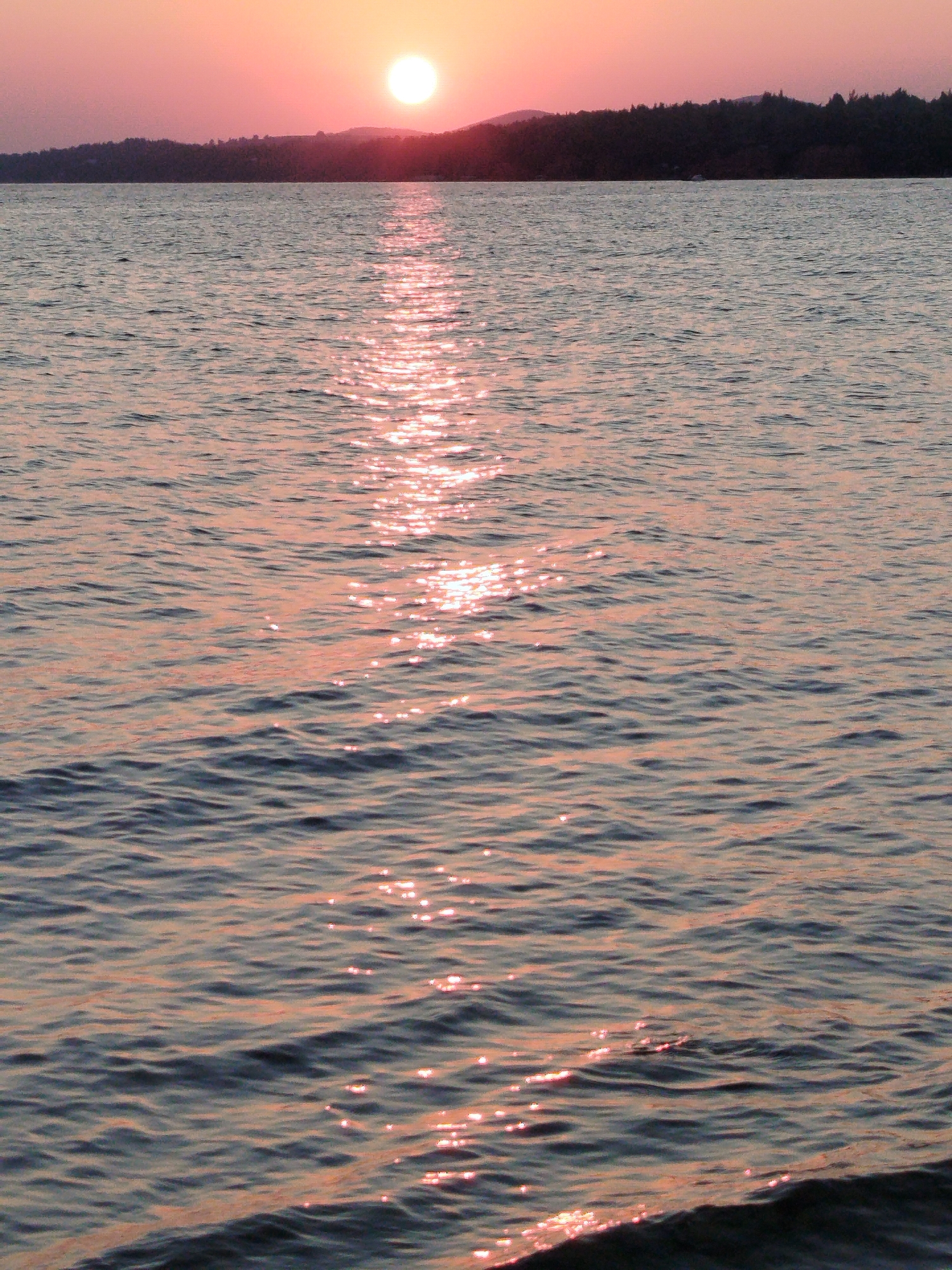 The image depicts a sunset over the sea in the Sithonia Municipal Unit, Greece. The sun is a bright, round orb low on the horizon, casting a golden and pinkish glow across the sky and water. A dark, forested coastline forms the background, with gentle hills visible behind it. The sea is calm, with small waves reflecting the sunlight in a shimmering path that stretches from the horizon towards the foreground. The overall atmosphere is serene and peaceful. There are no people or specific man-made objects clearly visible, other than what may be a small boat in the distance on the water.