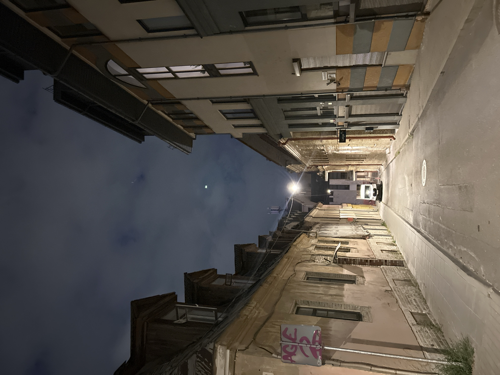 The image captures a narrow, dimly lit cobblestone street in Vilnius, Lithuania, at night. The sky is overcast and dark, with some faint cloud formations visible. The street is flanked by buildings of varying architectural styles and ages. On the left, older, weathered buildings with visible brickwork and sloping roofs are present, some with dormer windows. A sign with graffiti, reading "AGE 2", is mounted on a pole on the left side of the street.
On the right, a more modern building stands, featuring a cream-colored facade with horizontal bands of muted orange and grey. This building has several large, vertically oriented windows, some of which are illuminated from within. The ground floor of this building appears to have various entrances and shuttered windows.
In the distance, a white van is parked near the end of the street. Streetlights cast pools of light on the cobblestones and sidewalks, illuminating the scene. A few sparse plants grow along the edge of the sidewalk on the left. The overall atmosphere is quiet and somewhat eerie, typical of a city street at night. There are no people visible in the image.