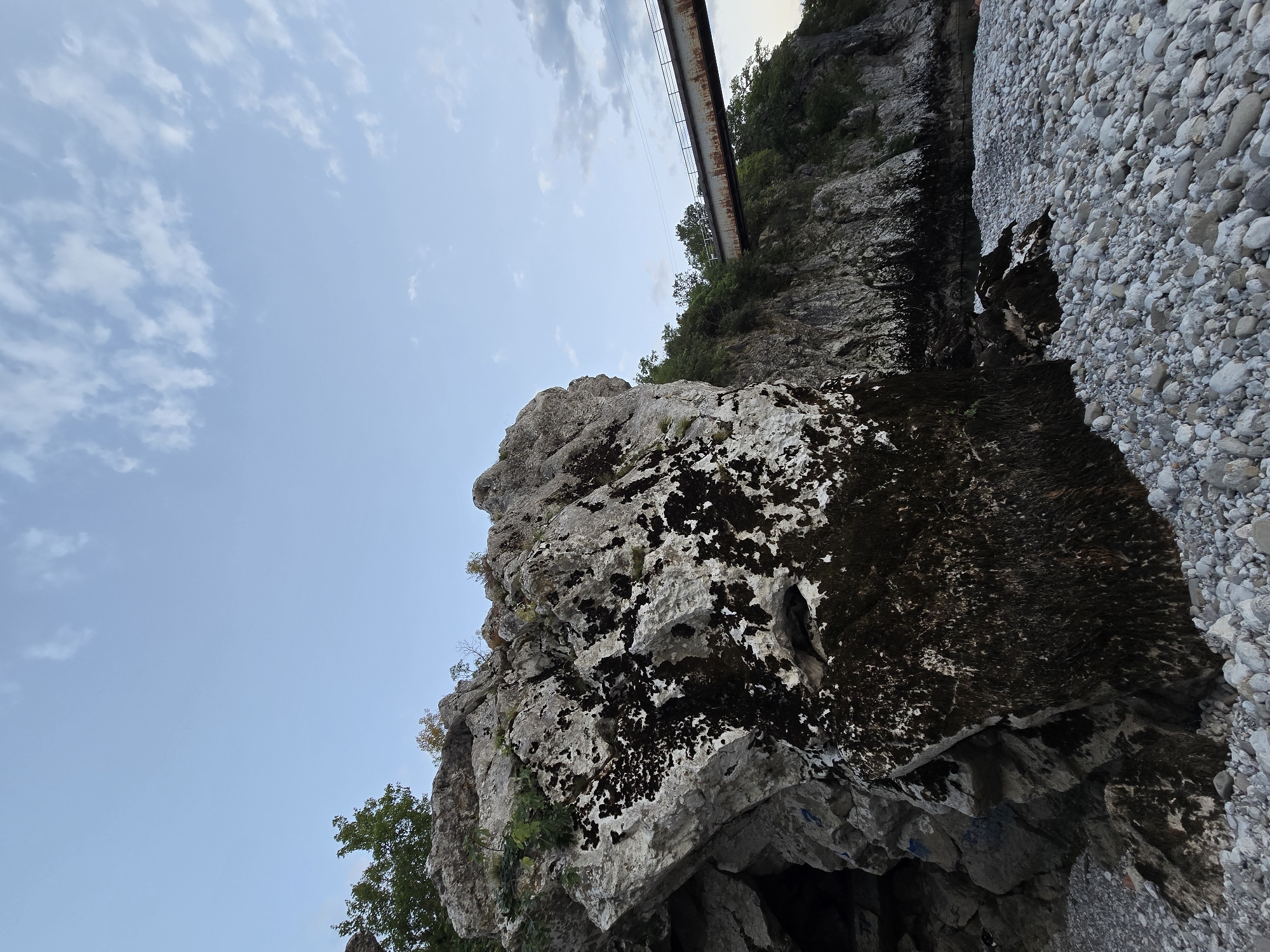 This is a vertical, wide-angle, eye-level shot of a rocky riverbed near a cliff face, with a bridge visible in the background. The riverbed is composed of smooth, white and grey pebbles. In the foreground, a large, dark, moss-covered rock juts out. Behind it, the cliff face rises, with patches of dark moss and some greenery. To the right, a rusty metal bridge spans the river, with trees lining the opposite bank. The sky is a pale blue with scattered white clouds. The scene appears to be in Bioče, Montenegro, judging by the presence of the bridge which is part of the Belgrade–Bar railway. No people are present.