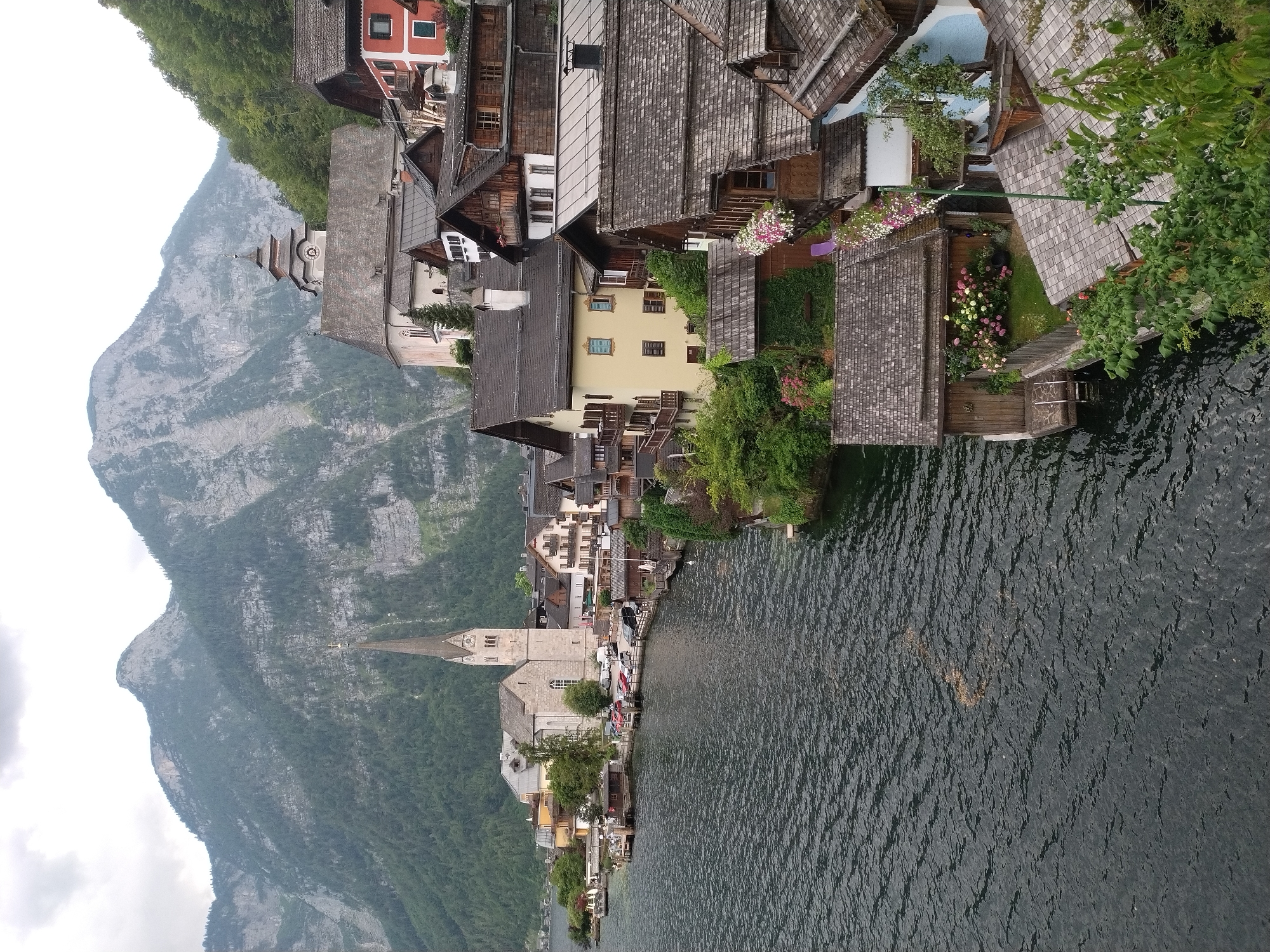 The image captures a picturesque view of Hallstatt, Austria, a lakeside village nestled against the backdrop of imposing mountains. The foreground is dominated by a dark, rippling lake, with a cluster of charming wooden houses built along its edge, some appearing to partially float on the water. The houses feature traditional architecture with steep, shingled roofs and wooden balconies. A notable church with a tall, slender spire and a clock face stands prominently in the midground, symbolizing the village's historic character. Beyond the village, steep, forested mountains rise sharply, their slopes covered in dense green foliage, creating a dramatic natural setting. The sky is overcast, casting a soft, diffused light over the scene. There are no visible people or specific actions, the image focuses on the static beauty of the landscape and architecture.