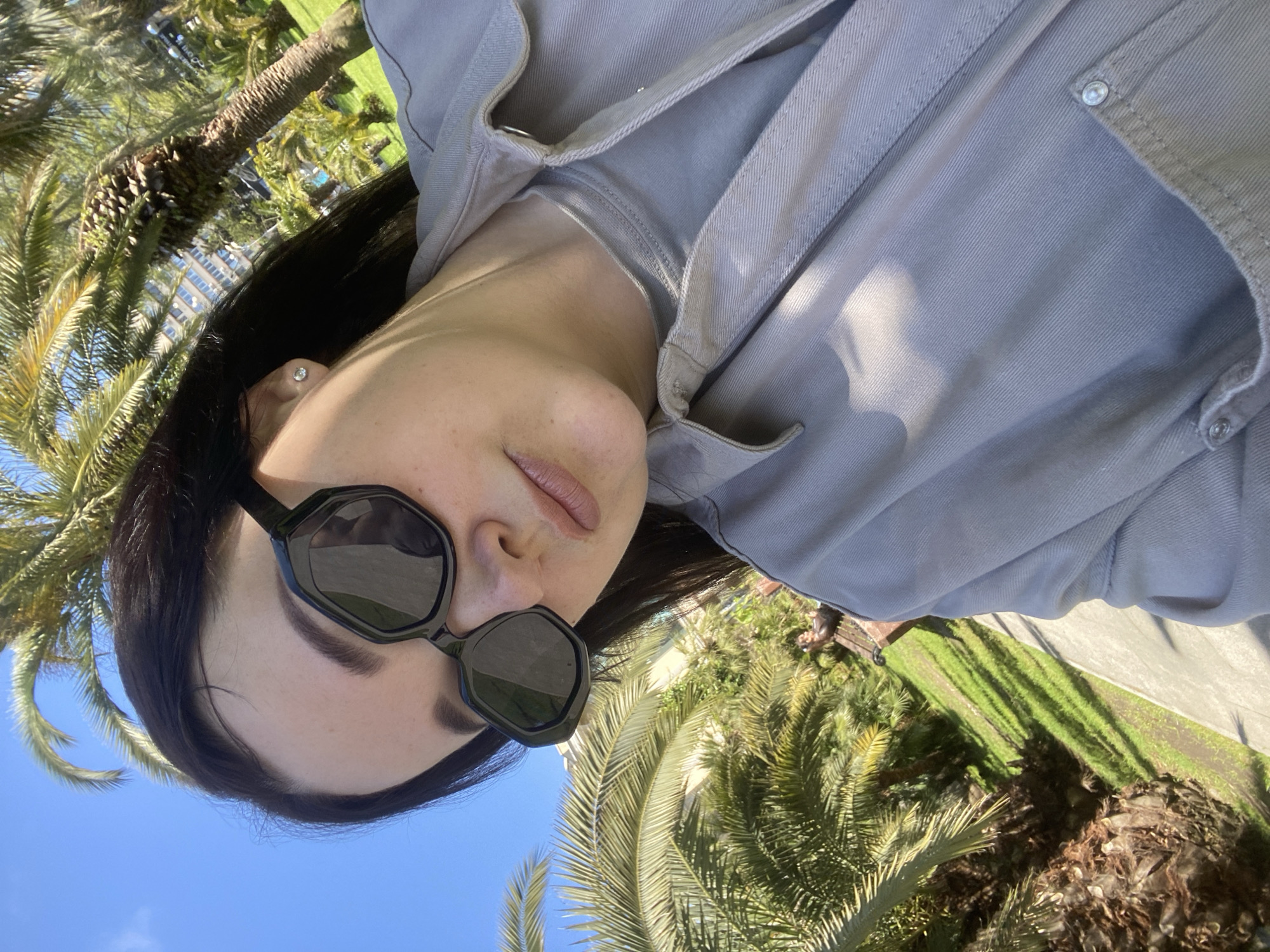 A woman is photographed in an outdoor setting with palm trees. She is wearing dark sunglasses and a button-down shirt. Lush green foliage is visible in the background along with the blue sky. Given the presence of palm trees and the city context of Sochi, Russia, the setting is likely a park or coastal area.