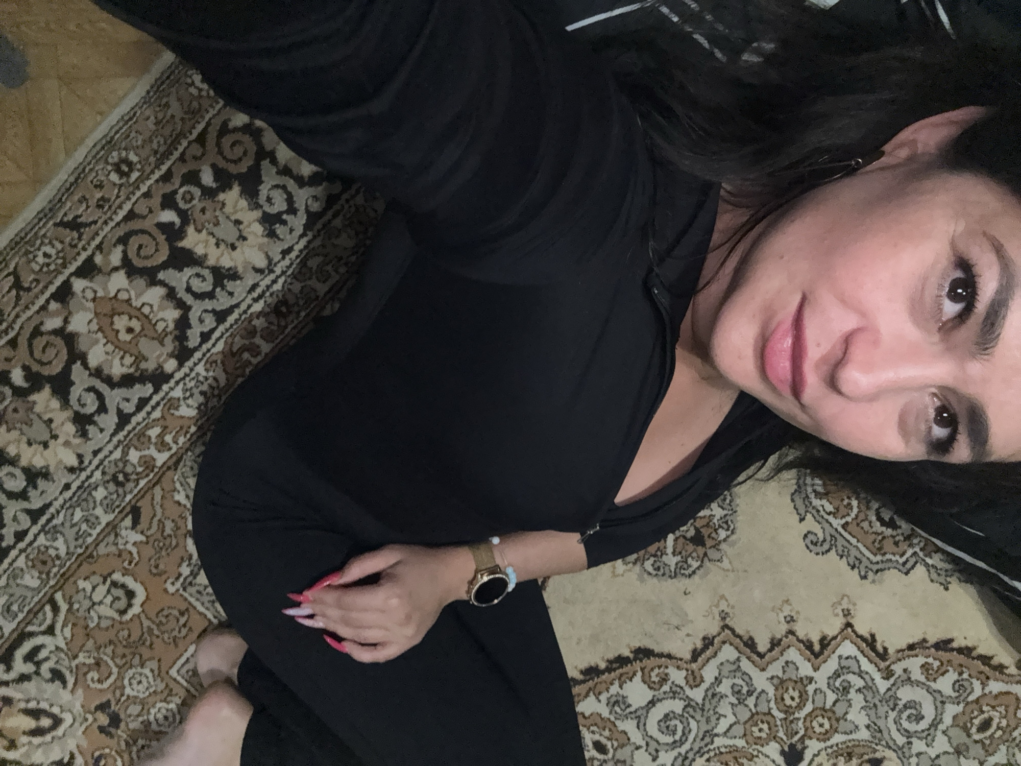 An indoor selfie from Chaikovsky, Russia, features a woman looking directly at the camera with a soft, slightly smiling expression. She is positioned on a floor covered by a traditional patterned rug, showcasing intricate designs in shades of beige, brown, black, and white.
The woman has long, dark hair and is wearing a black long-sleeved top and black pants. On her left wrist, she wears a gold-toned smartwatch with a black circular face, alongside a light-colored beaded bracelet. Her fingernails are long and manicured with a pink and white design. Her bare feet are partially visible at the bottom left of the frame. The lighting suggests an indoor setting, likely a residence. No specific time of day or external weather is discernible, and no text is visible within the image.