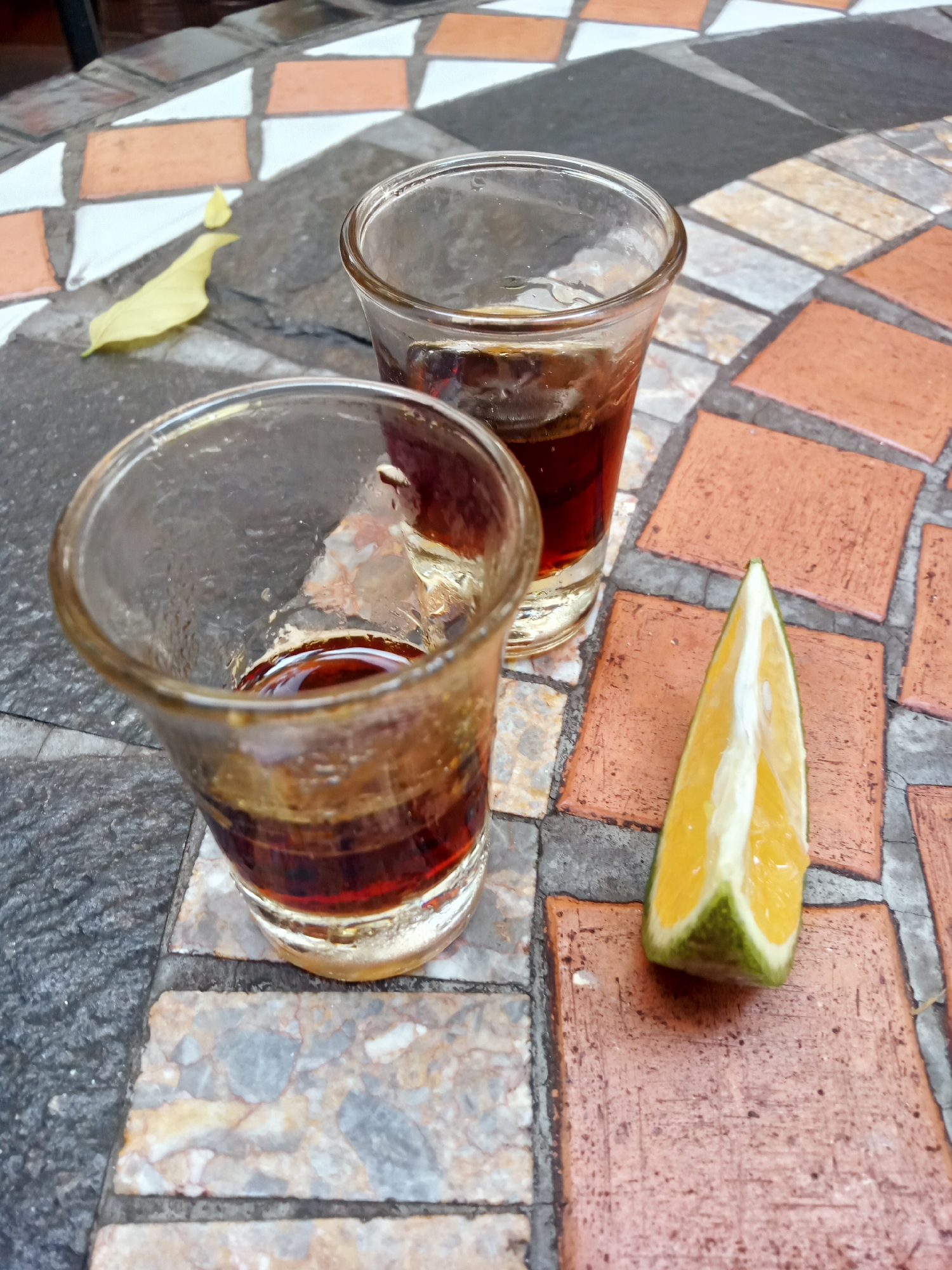 Two shot glasses containing a dark brown liquid sit on a mosaic tile table. Next to them is a wedge of citrus fruit with a green rind and yellow segments. A yellow leaf lies on the table in the upper left corner. The photo was taken in Nha Trang, Vietnam.