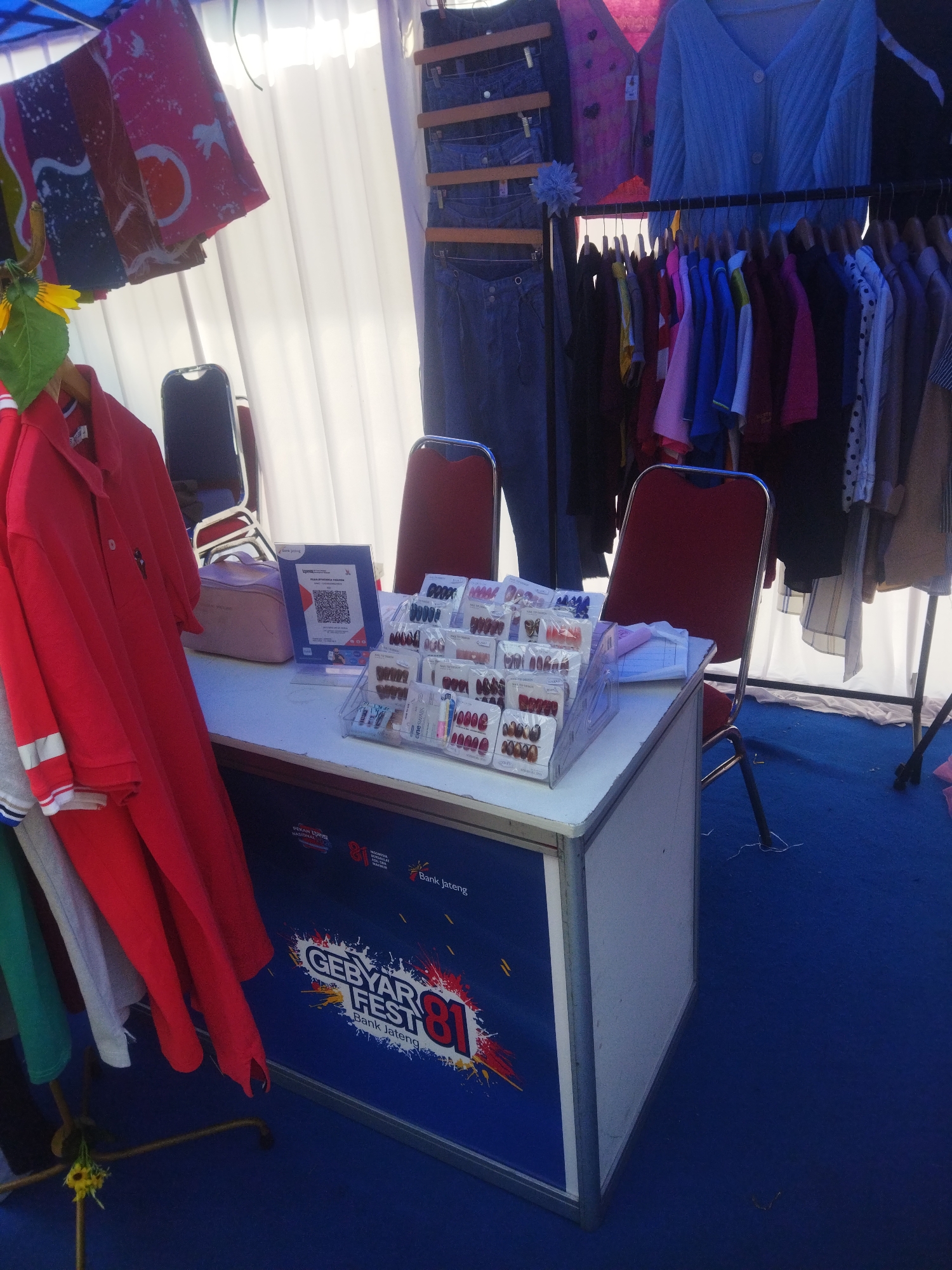 This image depicts an outdoor stall, likely part of a festival or market, set up on a vibrant blue carpet in the city of Wonogiri, Indonesia. The stall features a white table displaying various items, including rows of packaged false fingernails in clear plastic containers, a pink makeup bag, and a QR code for payment. Behind the table, several chairs with red upholstery and metal frames are visible. Hanging from racks, a collection of clothing items is displayed, including jeans, a blue knitted cardigan, colorful t-shirts, and patterned fabrics. A large blue banner with white text and graphics, featuring the words "GEBYAR FEST 81" and "Bank Jateng," adorns the front of the stall. The background consists of white curtains and a clothing rack. The overall scene suggests a commercial or promotional event, with an emphasis on retail goods.
