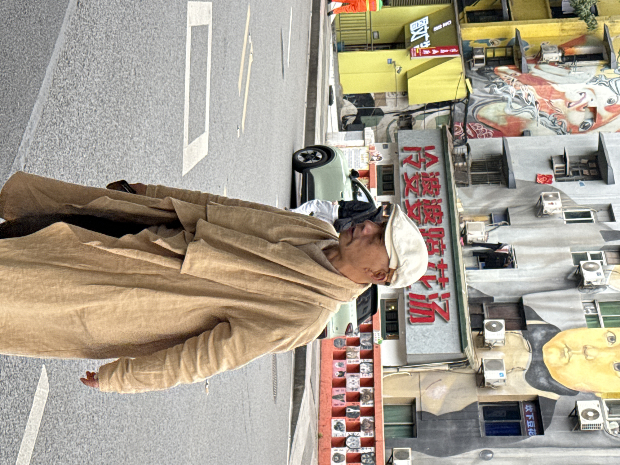 An adult male, positioned right of center, walks left-to-right across a paved road surface. He wears a light beige flat cap, a long, layered, light brown or beige coat, and a draped scarf or cowl of the same color. His left hand holds a dark-colored smartphone.
The scene is an urban street in Jiulongpo District, China. The background features multi-story buildings adorned with large, colorful murals and numerous external air conditioning units. The building on the left is primarily yellow with a vibrant mural depicting a stylized face or creature. The adjacent buildings are gray, one featuring a prominent mural of a yellow-skinned baby or child's face, and another with a large red sign displaying Chinese characters "冷婆婆辣花汤" (Leng Po Po La Hua Tang, meaning "Granny Leng's Spicy Flower Soup"). A smaller blue sign reads "炊下豆花" (Chui Xia Dou Hua, meaning "Cooked Bean Curd"). A person is visible on an upper-level balcony or window, dressed in dark clothing.
On the road, white painted markings are visible, including a rectangular box and two parallel short lines. A light green compact SUV is situated behind the main subject, on the right side of the road, with a partially visible person in dark clothing and white pants near it. To the far left, a person in a bright orange jumpsuit walks on the sidewalk. A low, orange concrete barrier or wall on the right features multiple recessed squares, each containing a painted image of a dog's face.