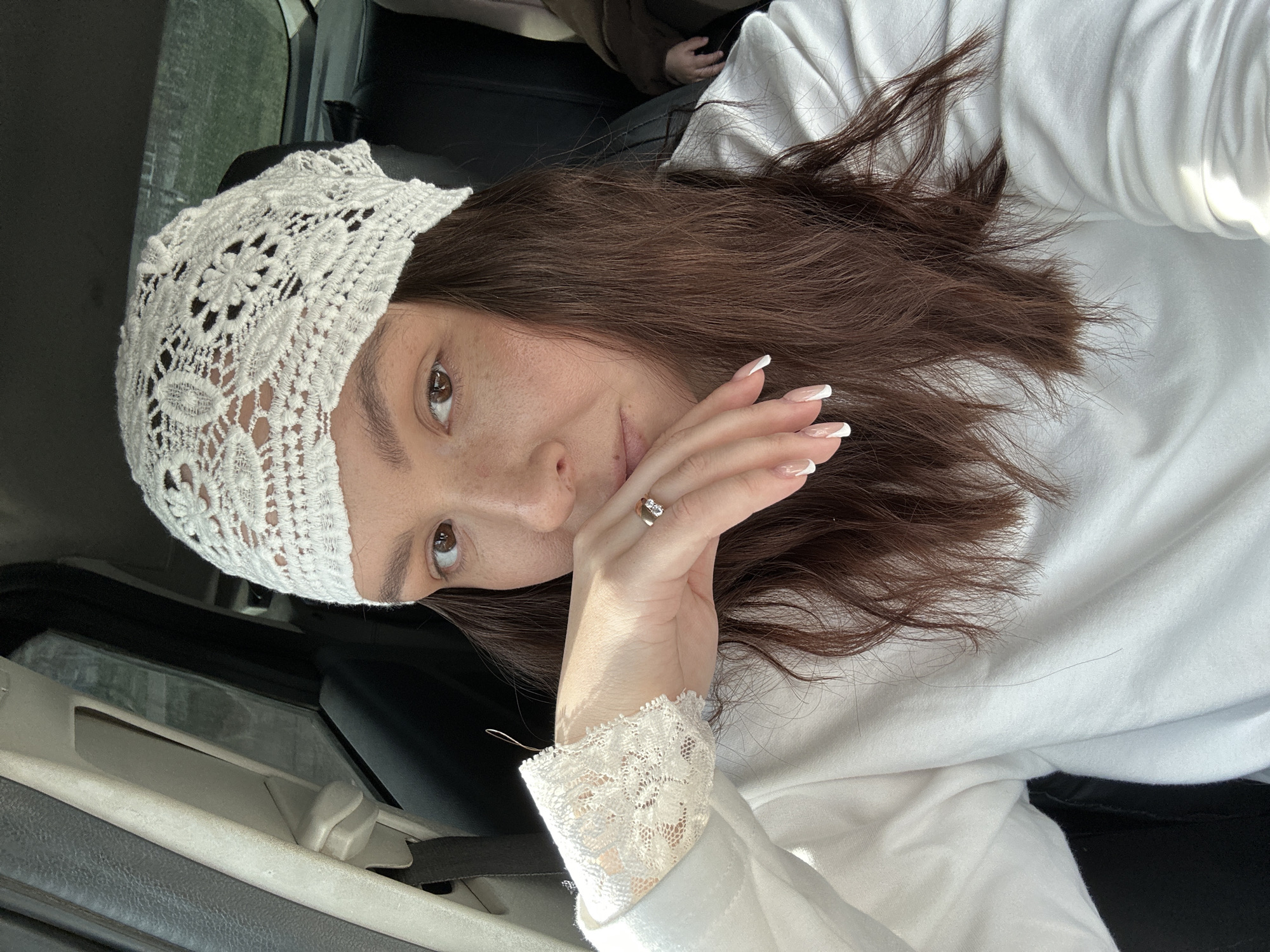A woman with brown hair and hazel eyes is wearing a white lace head covering and a white long-sleeved shirt with a lace cuff. She is holding her hand near her face, and a ring is visible on her finger. The background suggests she is inside a vehicle, possibly a car, with elements of the car interior visible. The image appears to have been taken with a mobile phone. There is no verifiable evidence to support the claim that this image was taken in Noviy, Russia. Therefore, this detail is omitted.