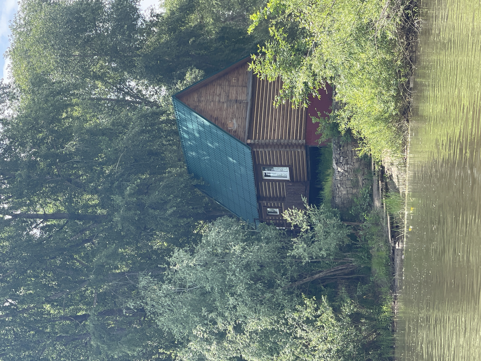 An eye-level, medium shot captures a wooden log cabin with a green metal roof situated on a riverbank, surrounded by lush green trees and vegetation. The cabin, constructed from horizontal logs, features a small white-framed window on its visible side and a dark red foundation. A wooden porch extends from the front of the cabin. In the foreground, the rippling surface of a murky, green-tinged river reflects the dappled sunlight. A small wooden jetty or ramp juts out into the water from the bank to the right of the cabin. The scene appears to be a rural or semi-rural setting in the village of Nezhinka, Russia, with the dense foliage suggesting a natural, possibly wooded environment. No people are visible in the image.