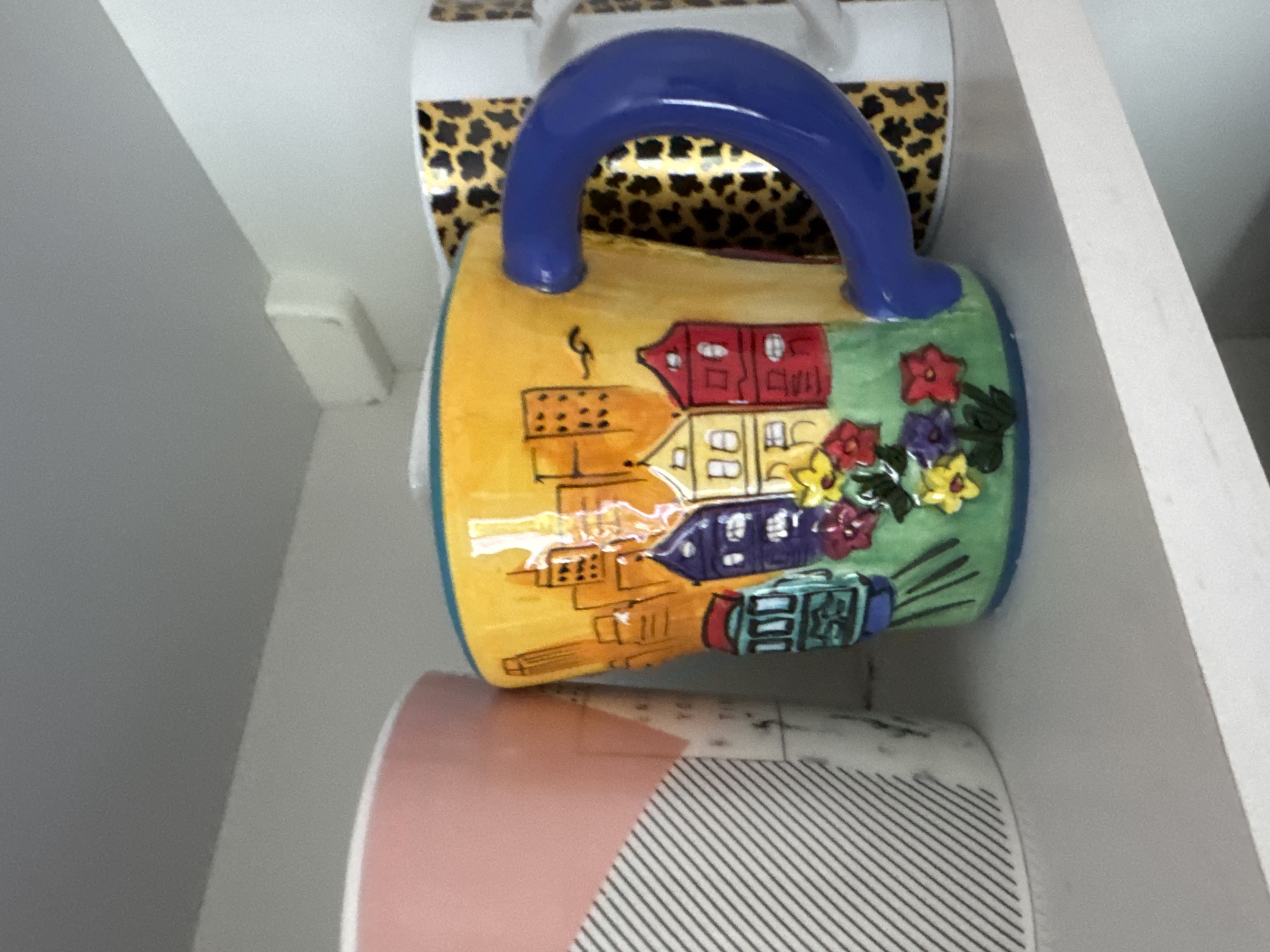 The image shows three mugs on a white shelf. The mug in the foreground is yellow and decorated with a scene of a city skyline with colorful buildings and a tram. It has a large blue handle. To the left of this mug is another mug with a pink and white striped pattern. To the right, partially visible, is a mug with a leopard print pattern. The background appears to be a white wall with a light fixture. There is no indication of the location being Höhenkirchen-Siegertsbrunn, Germany.