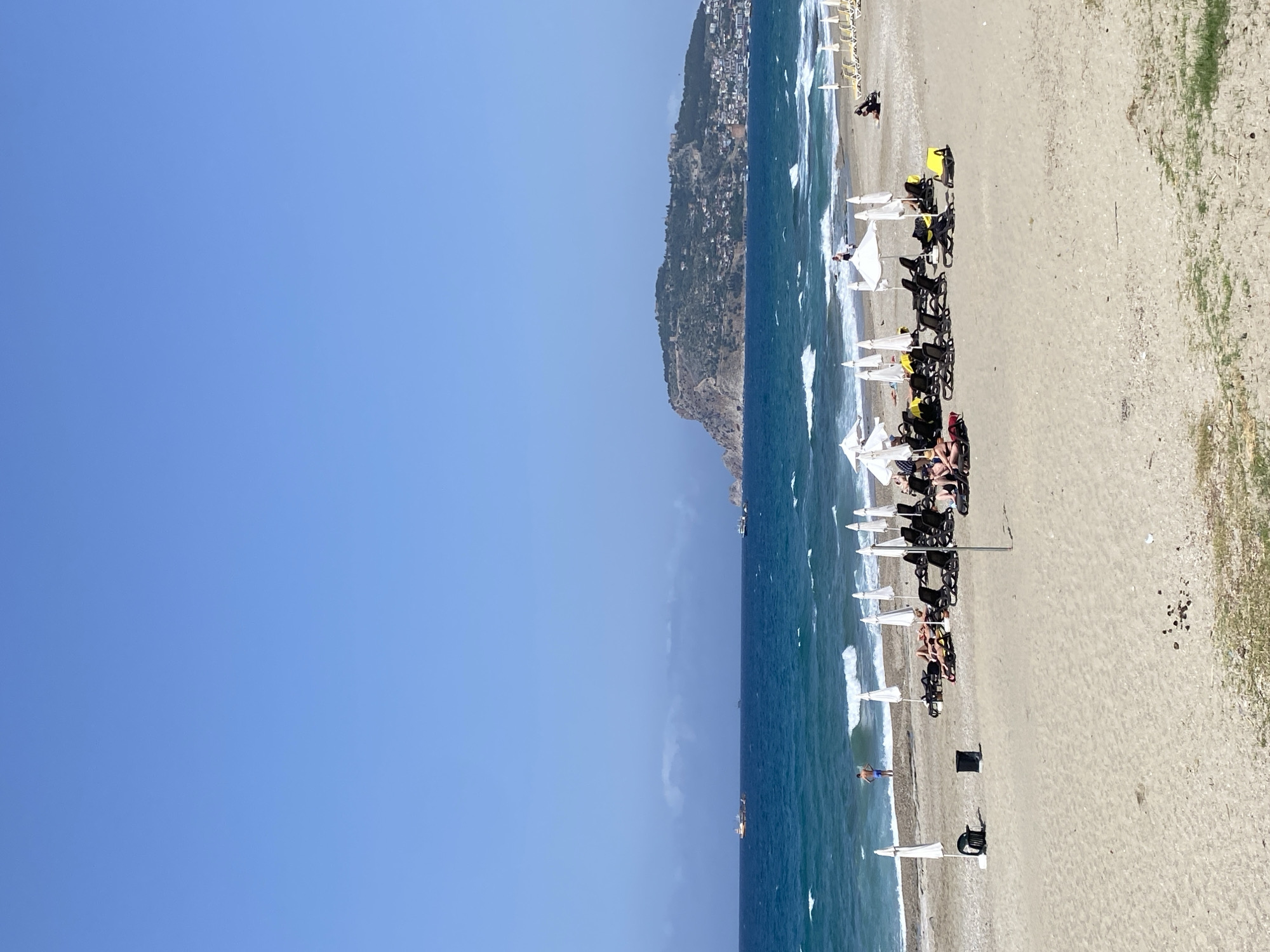 The image shows a beach in Alanya, Turkey. The sky is clear and blue, occupying about half of the image. The sea is a deep blue with white waves crashing on the shore. A cliff with buildings visible is on the horizon. A line of white beach umbrellas and chairs stretches along the sandy beach. People are lounging under some of the umbrellas. Some beach chairs are yellow. Some people are visible in the water.