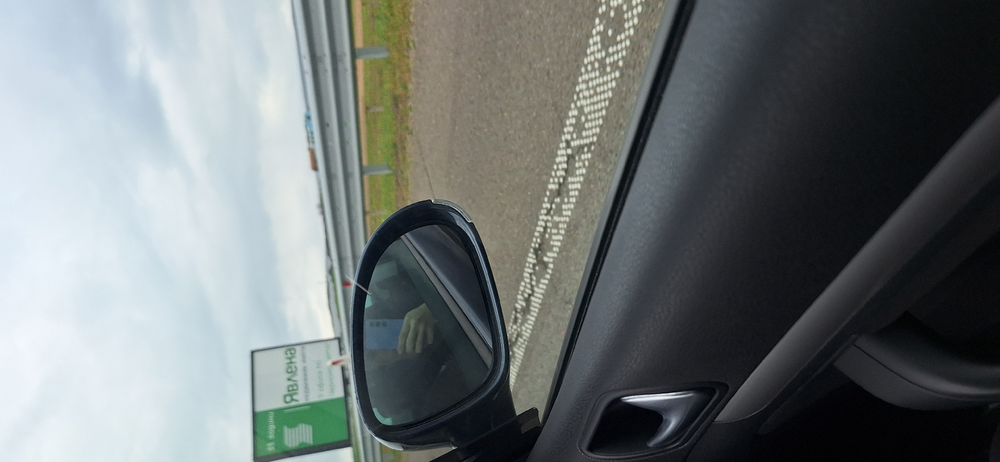 The image, taken from the interior of a moving vehicle, shows a side view mirror reflecting a road sign for "Явлена" in Vratitsa, Bulgaria. The sign is green and white with text in Cyrillic. The side view mirror is black. Part of the driver's hand is visible in the side view mirror holding a blue phone. The road outside has a dotted white line along the edge. A metal guardrail is visible beyond the road. The sky is overcast. The car interior is mostly black and gray.