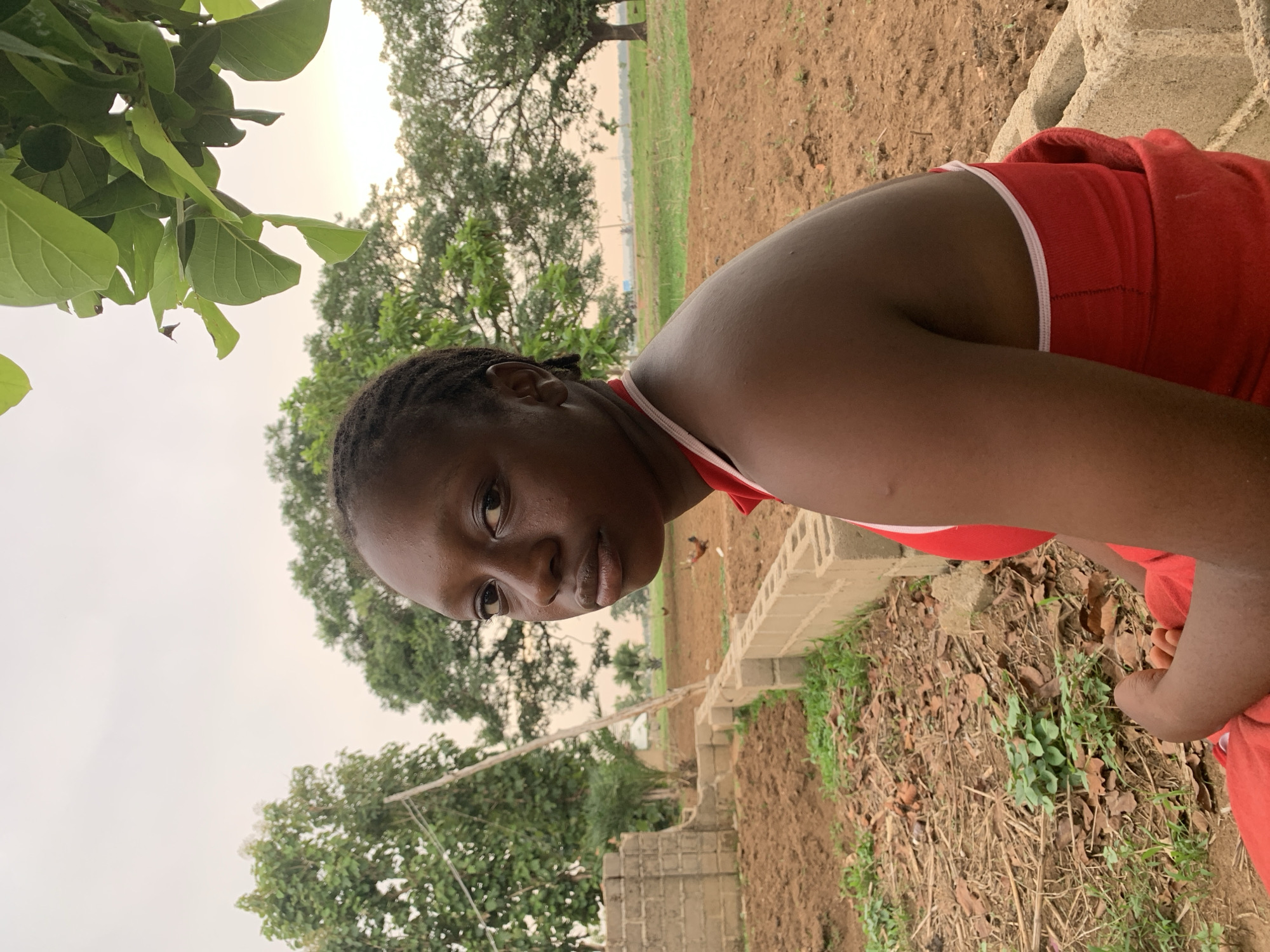 A woman in a red shirt is outdoors in Agban, Nigeria. She is partially obscured, with the focus on her face and torso. Behind her is a landscape with trees, a field, and a building structure made of blocks. The ground is dirt and foliage. A chicken can be seen behind her head to the right. Green leaves hang over the scene from the left, suggesting proximity to a tree. The sky is overcast.