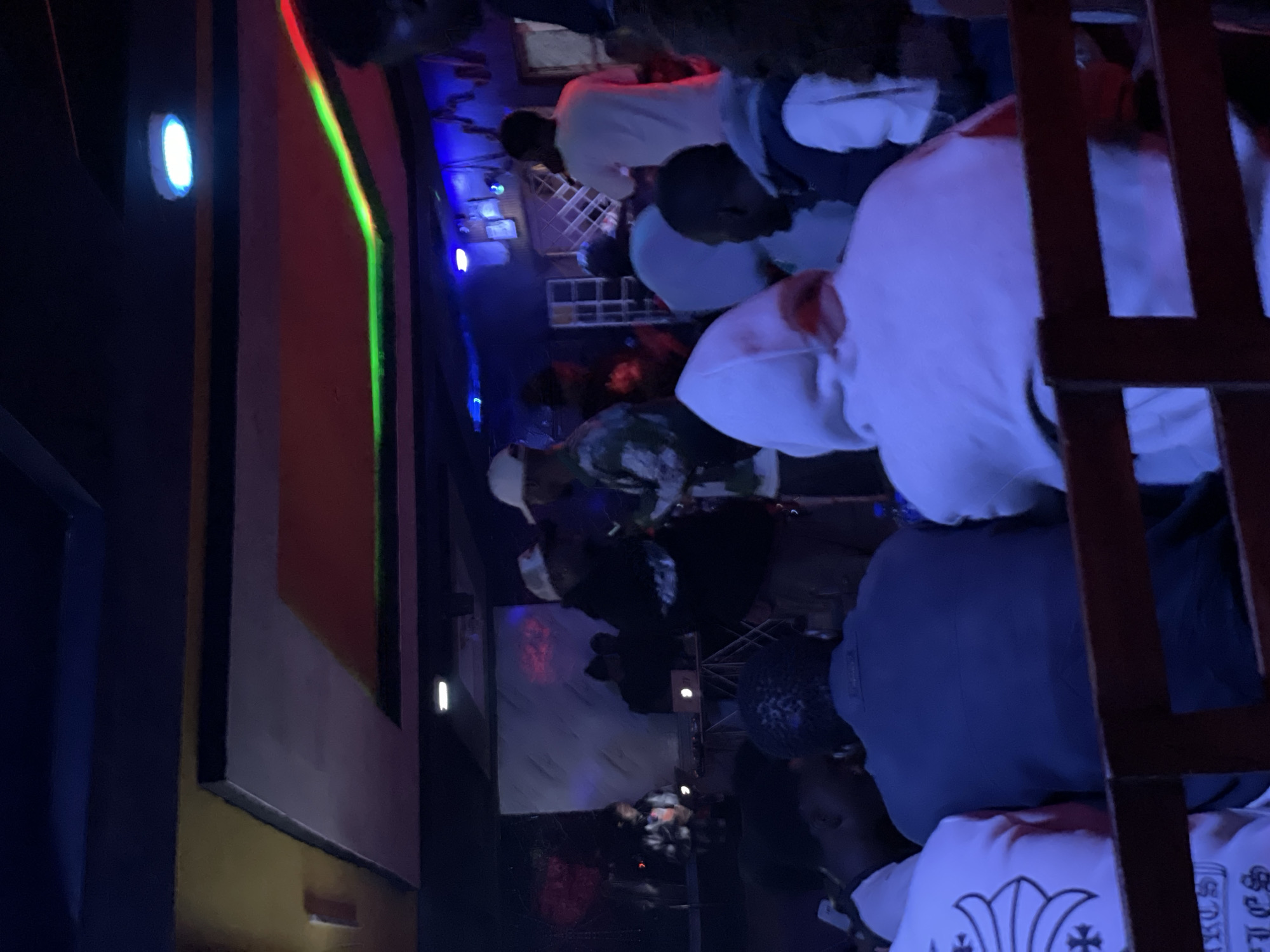 The media file captures a dimly lit indoor scene, likely a social gathering or party in Durumi, Nigeria. Several individuals are visible, predominantly wearing casual clothing such as hoodies and t-shirts. The lighting is atmospheric, with colorful neon lights—including red, green, and blue—casting shadows and illuminating parts of the scene. The presence of what appears to be a bar or serving area with bottles and glasses suggests a recreational setting. Some individuals are standing, while others are seated or leaning, engaged in conversation or observation. The overall mood seems relaxed and social. The time of day is implied to be evening or night due to the artificial lighting and the atmosphere. No specific weather conditions are discernible as the scene is indoors. There are no immediately obvious signs indicating the exact location beyond the provided context of Durumi, Nigeria.