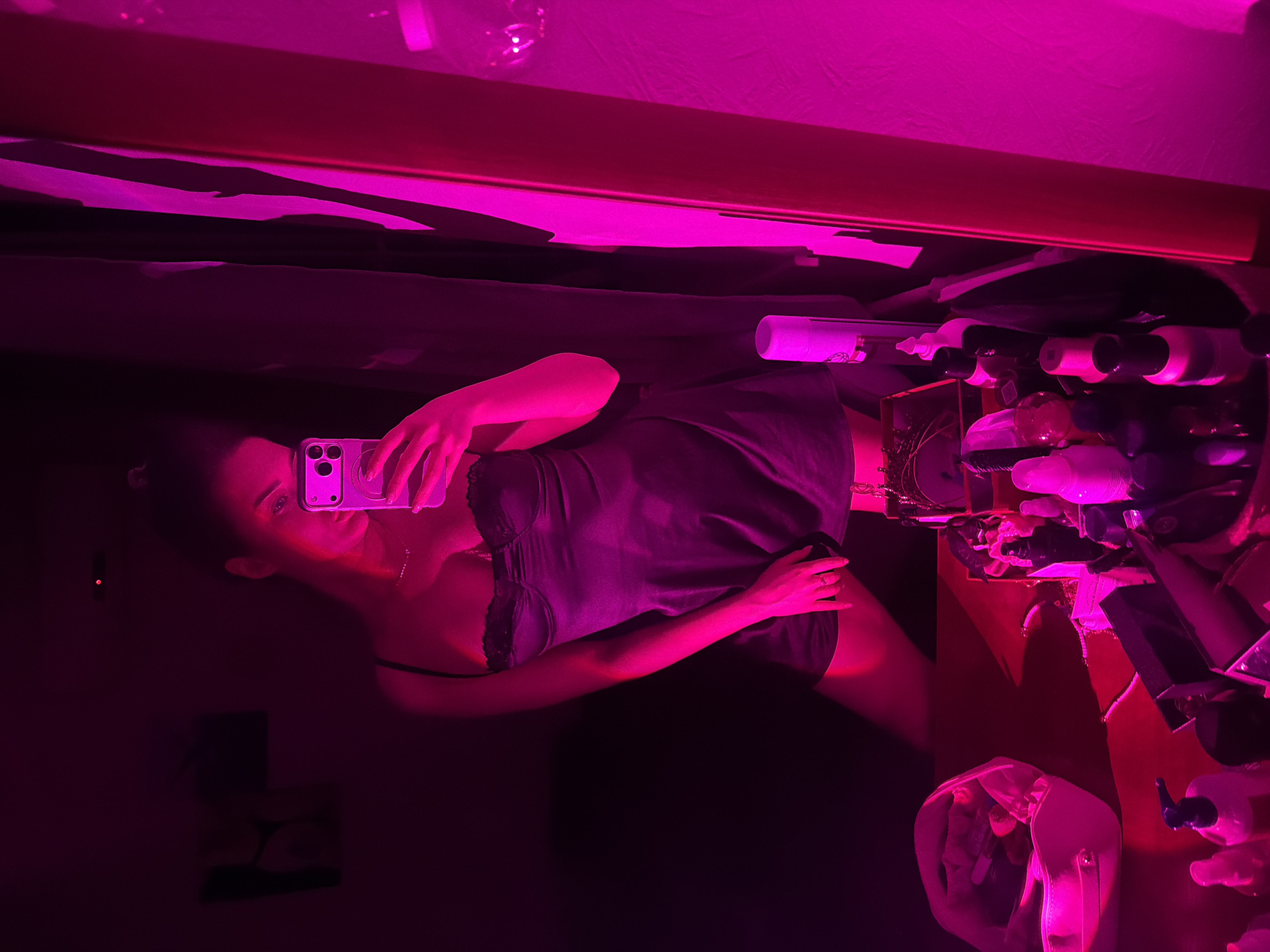A young woman is taking a selfie in a room lit with bright pink light. She is wearing a dark negligee with lace trim. The background is cluttered with various items on a table or dresser, including bottles, containers, and a clear bag. The reflection of the room's ceiling and other objects can be seen in a dark surface, likely a mirror or window, behind the woman. The location and whether it is in Moscow, Russia, cannot be determined from the image alone.